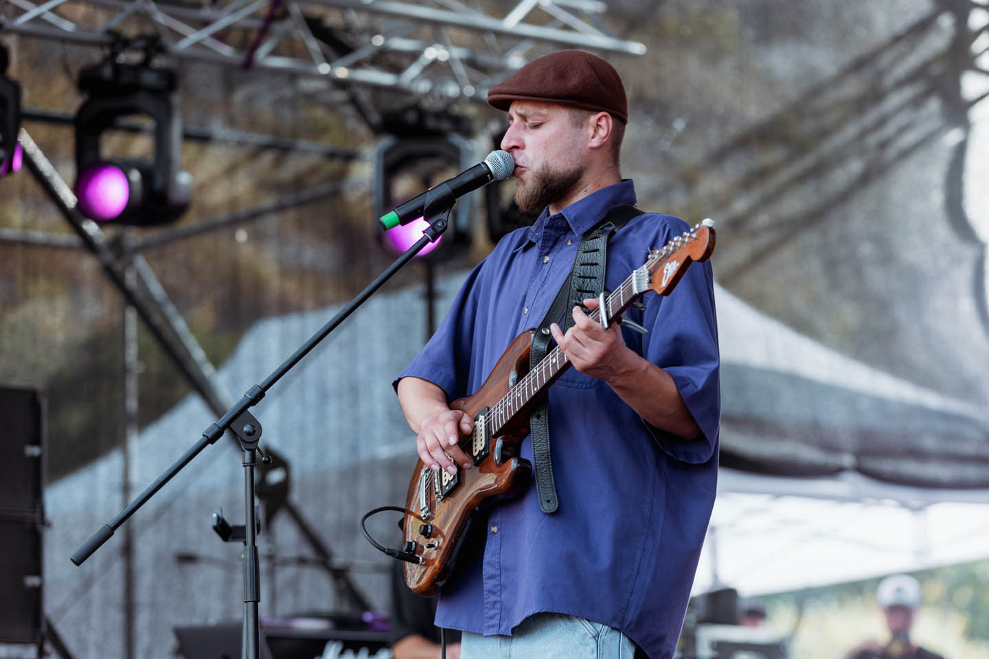Музыкант в коричневой шапке и синей рубашке поет в микрофон и играет на электрогитаре на сцене.