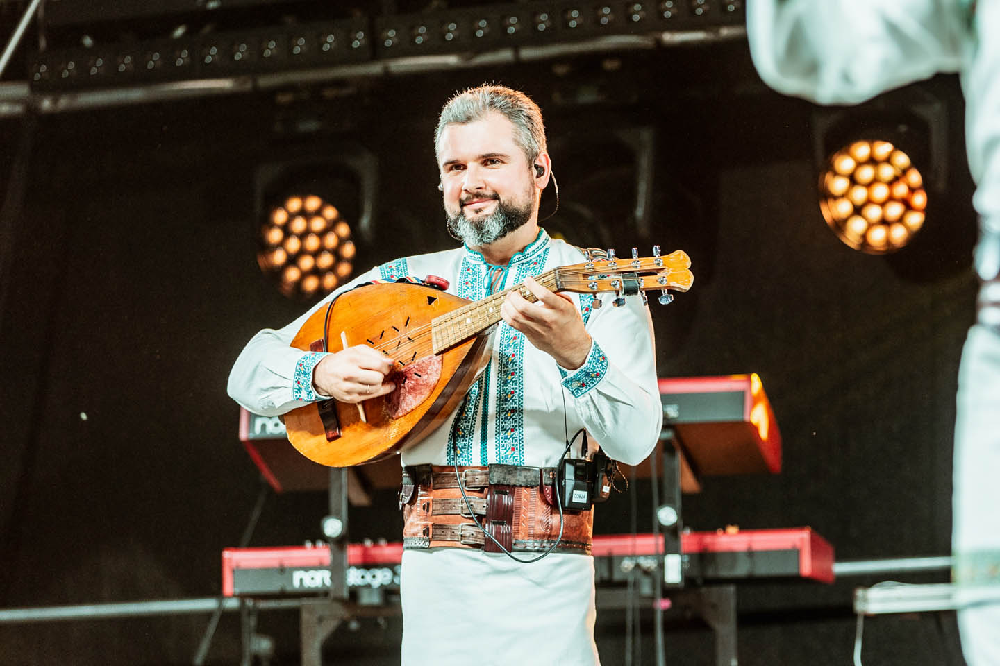 Музыкант в традиционной вышиванке и кожаном ремне играет на деревянном струнном инструменте на сцене.