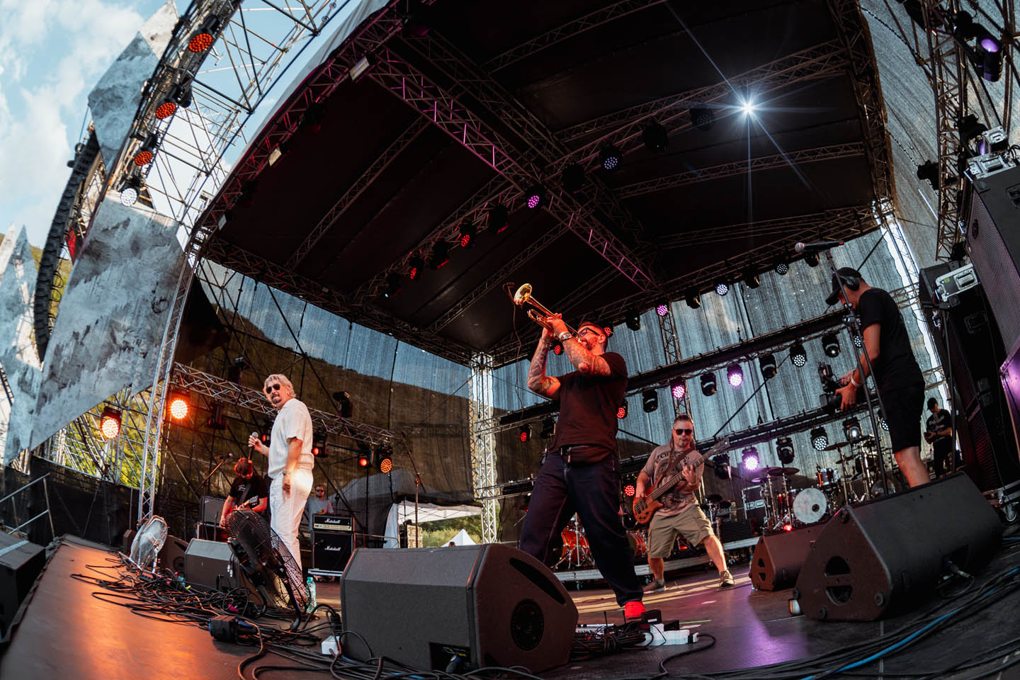 Живая группа музыкантов на сцене под открытым небом с трубачом, вокалистом и гитаристом во время концерта.