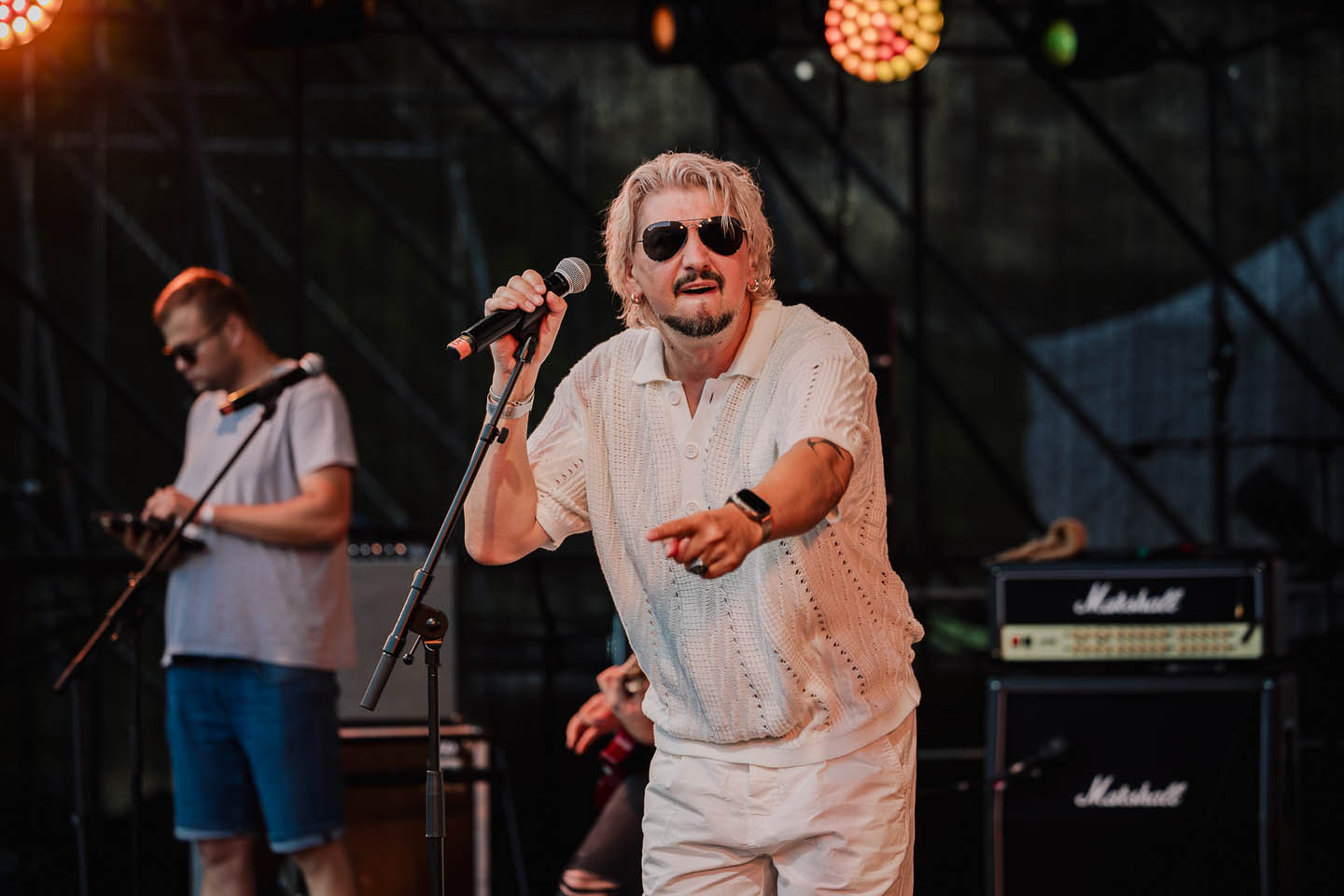 Исполнитель с светлыми волосами и темными очками, держащий микрофон и указывающий вперёд на сцене.