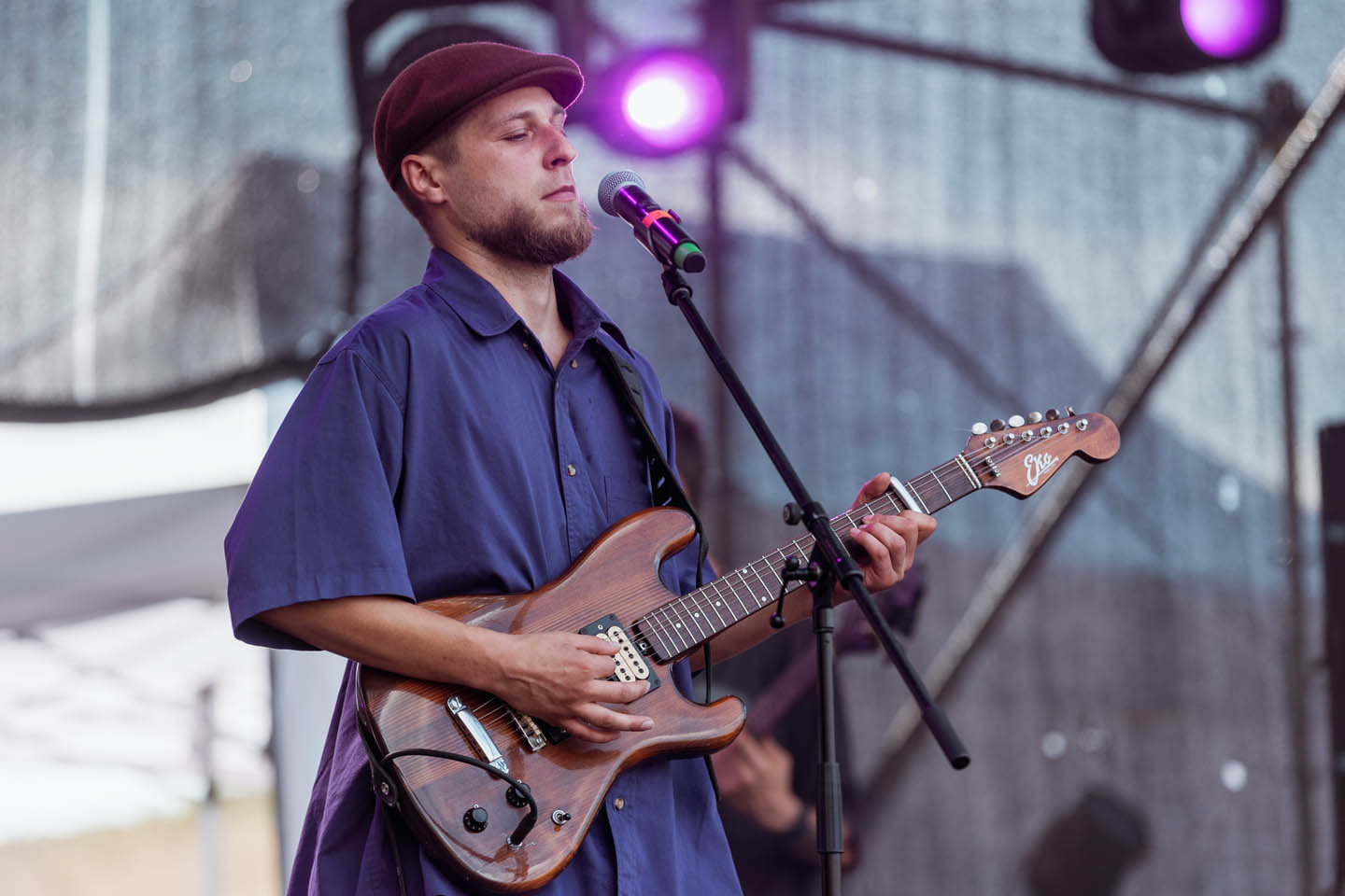Музыкант в бордовой кепке и синей рубашке играет на электрогитаре на сцене с микрофоном.