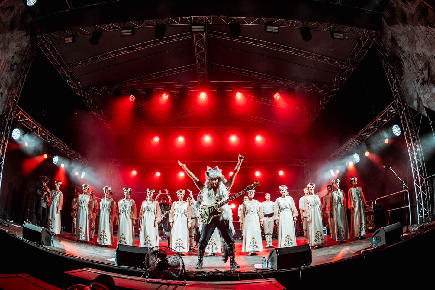 Исполнители на сцене с красным освещением, один человек в костюме волка с бас-гитарой, остальные в белых платьях с цветочной вышивкой.