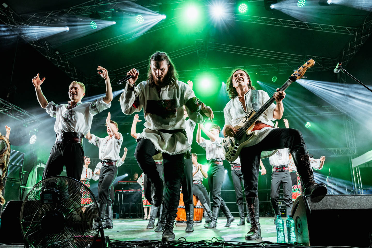 Группа музыкантов в традиционной одежде выступает на сцене с ярким зеленым освещением.