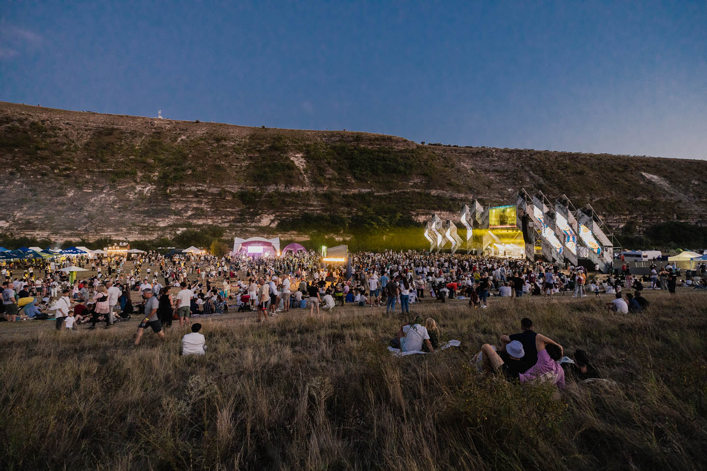 Большая толпа людей на открытом воздухе у сцены с подсветкой на фоне высокого холма в сумерках.