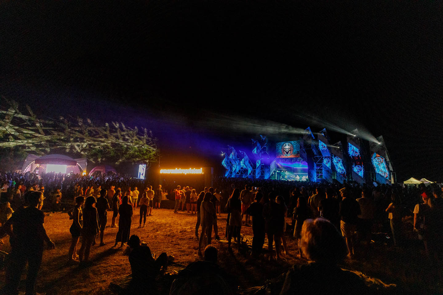 Ночная сцена на музыкальном фестивале с синими огнями и толпой людей, стоящих на открытом воздухе.