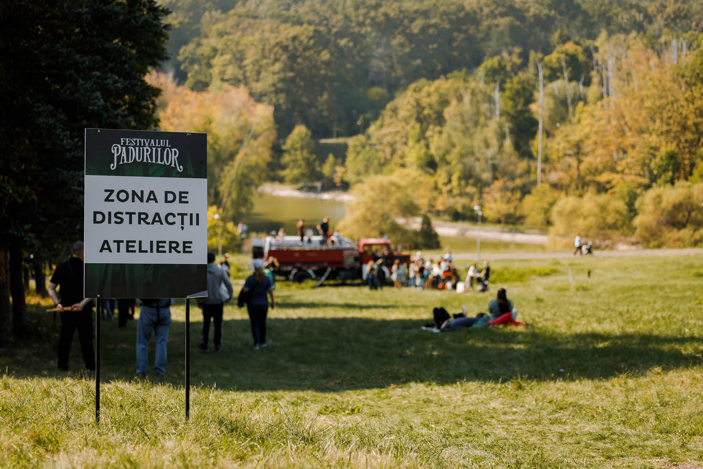 Люди на зелёной поляне в парке возле таблички «Зона развлечений и мастерских» на фестивале лесов.