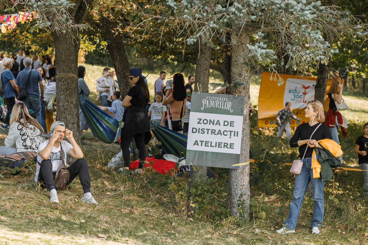 Люди отдыхают и общаются в лесном парке у таблички с надписью «Zona de Distracții Ateliere» на фестивале природы.