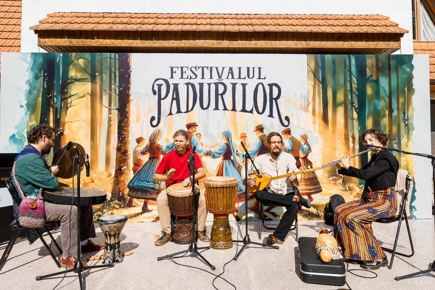 Четыре музыканта играют на традиционных инструментах перед ярким баннером с надписью «Festivalul Pădurilor» и изображением танцующих людей в народных костюмах на фоне леса.