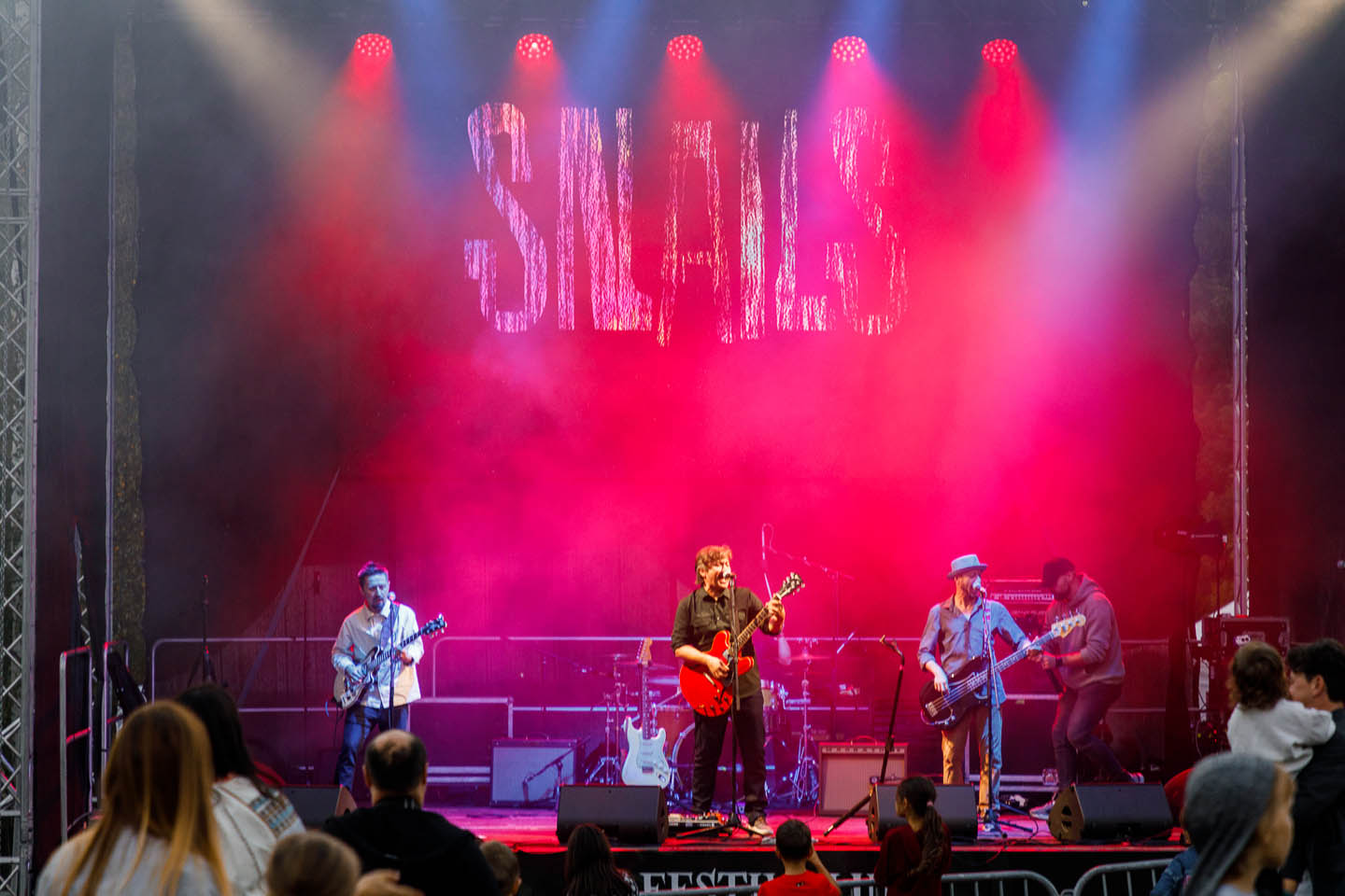 Группа музыкантов играет на гитарах и бас-гитаре на сцене с красным освещением и надписью SNails на заднике.