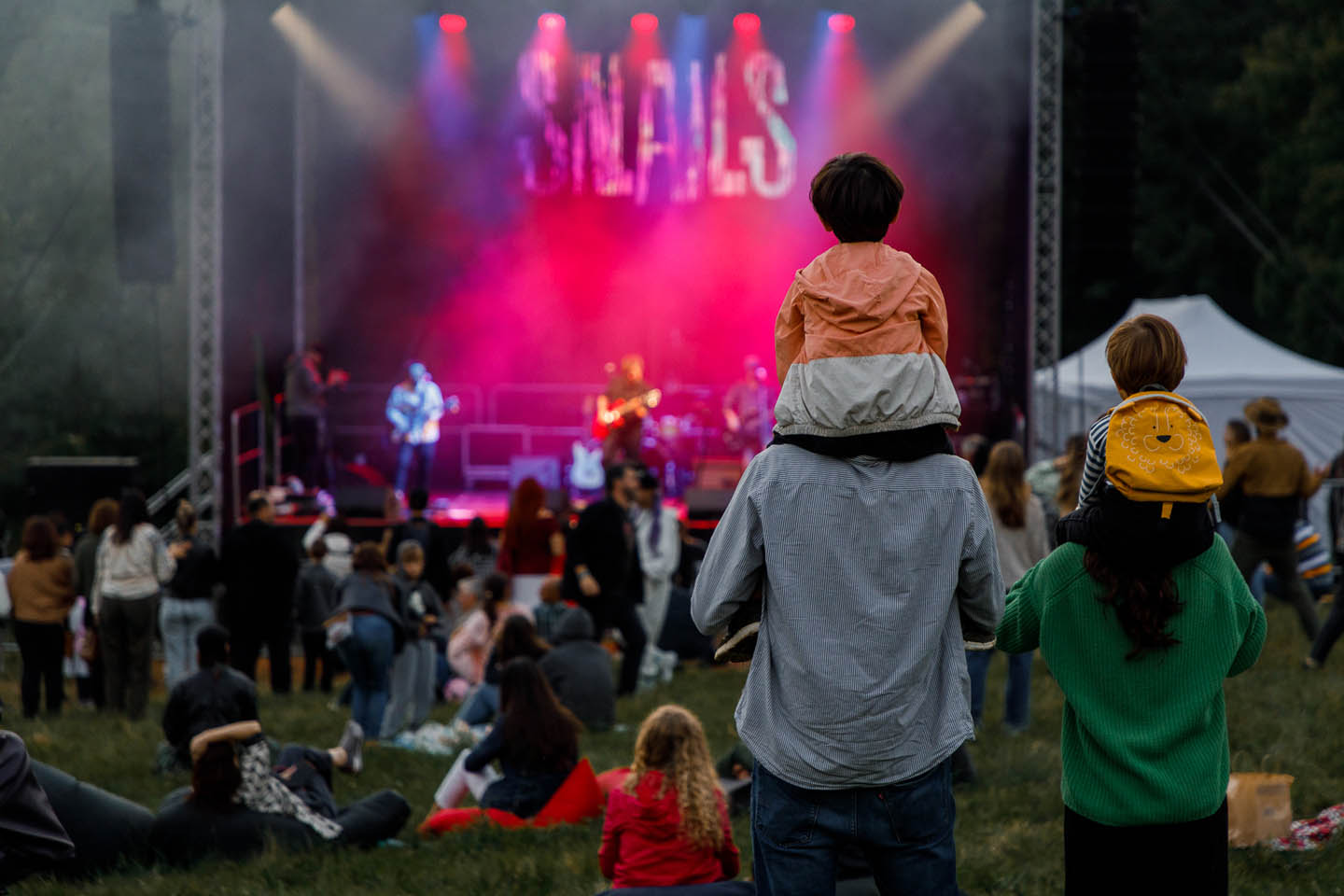 Группа людей смотрит на музыкальный концерт на открытом воздухе, двое детей сидят на плечах взрослых.