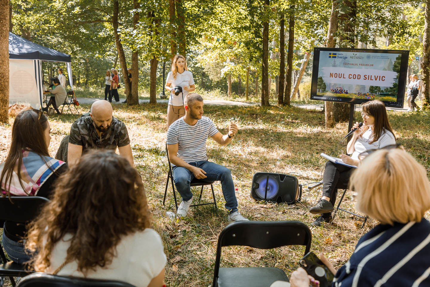 Группа людей на открытом воздухе участвует в дискуссии рядом с экраном с надписью «Noul Cod Silvic» в лесу.