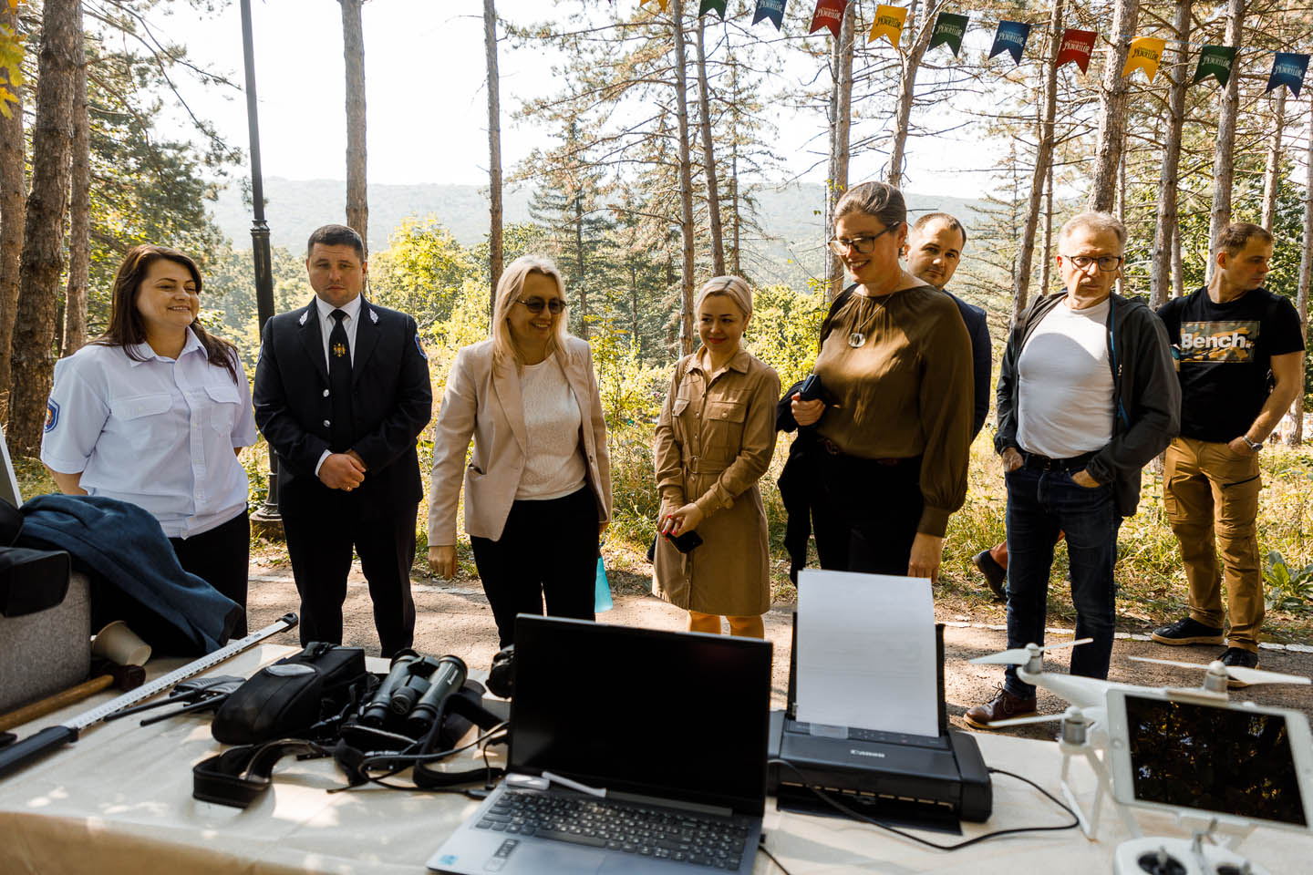 Группа людей стоит на открытом воздухе у стола с ноутбуком, принтером, биноклем и дроном на фоне леса.