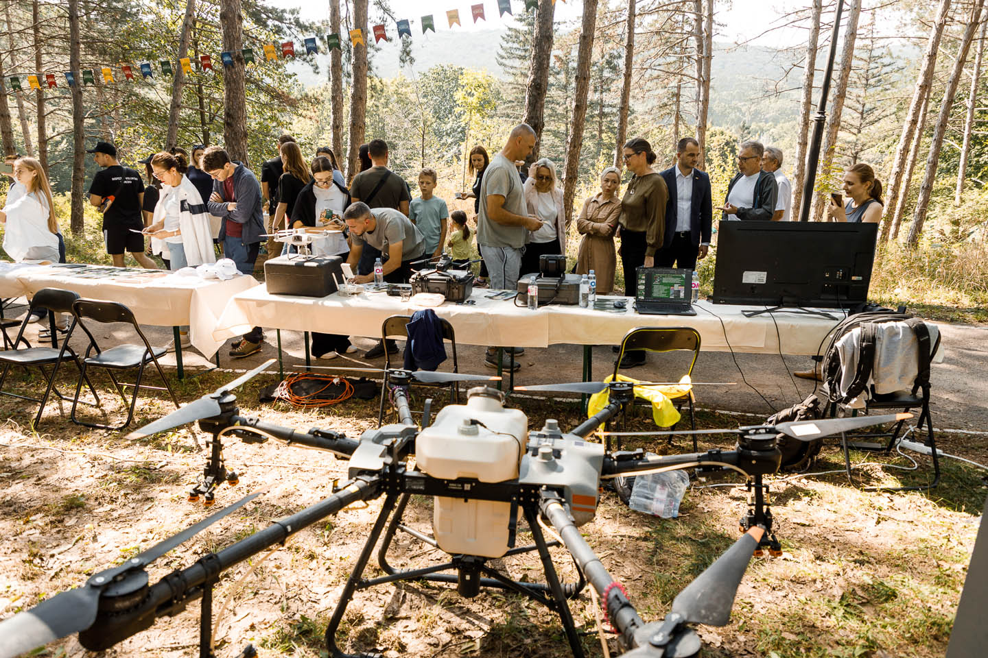 Группа людей на открытом воздухе изучает и обсуждает большой профессиональный дрон, установленный перед столом с оборудованием и электронной техникой в лесу.