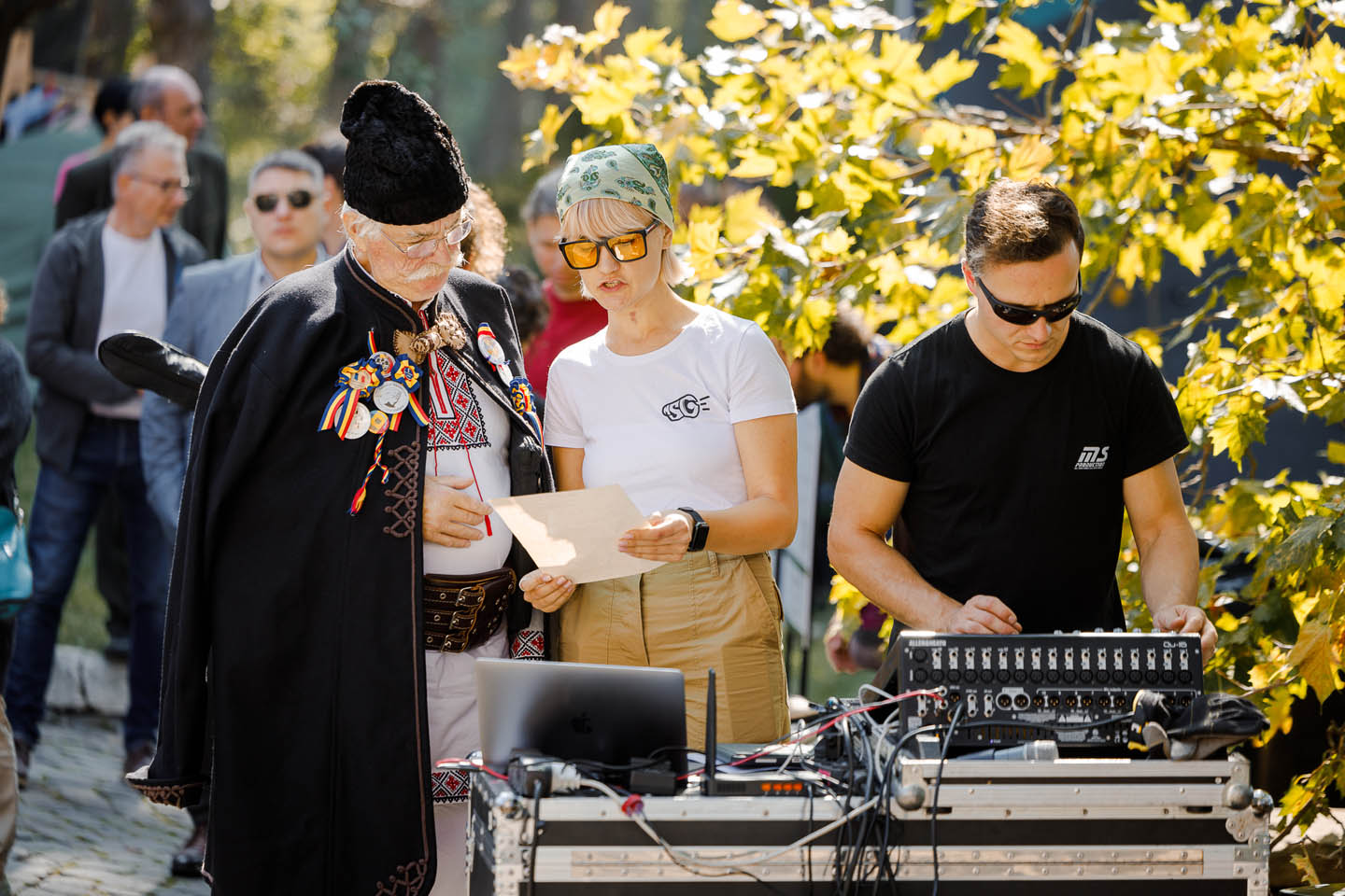 Трое людей на улице: пожилой мужчина в традиционной одежде с медалями, женщина с платком на голове и очками в белой футболке и мужчина в черной футболке с аудиоаппаратурой на столе.