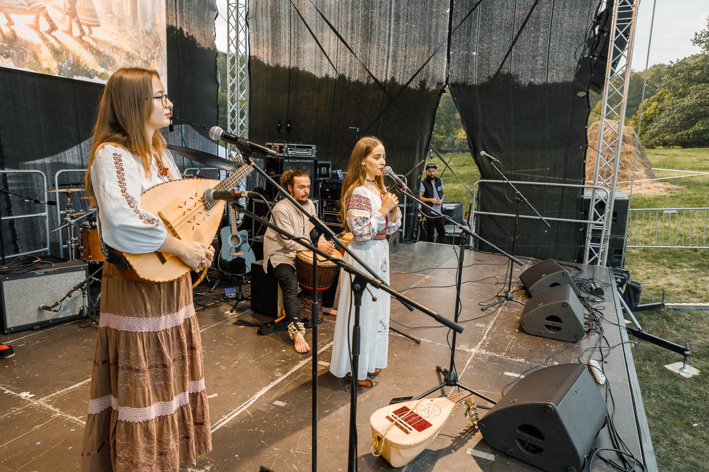 Музыкальный коллектив в национальных костюмах выступает на открытой сцене, одна девушка играет на бандуре, мужчина играет на барабане, другая девушка поет в микрофон.