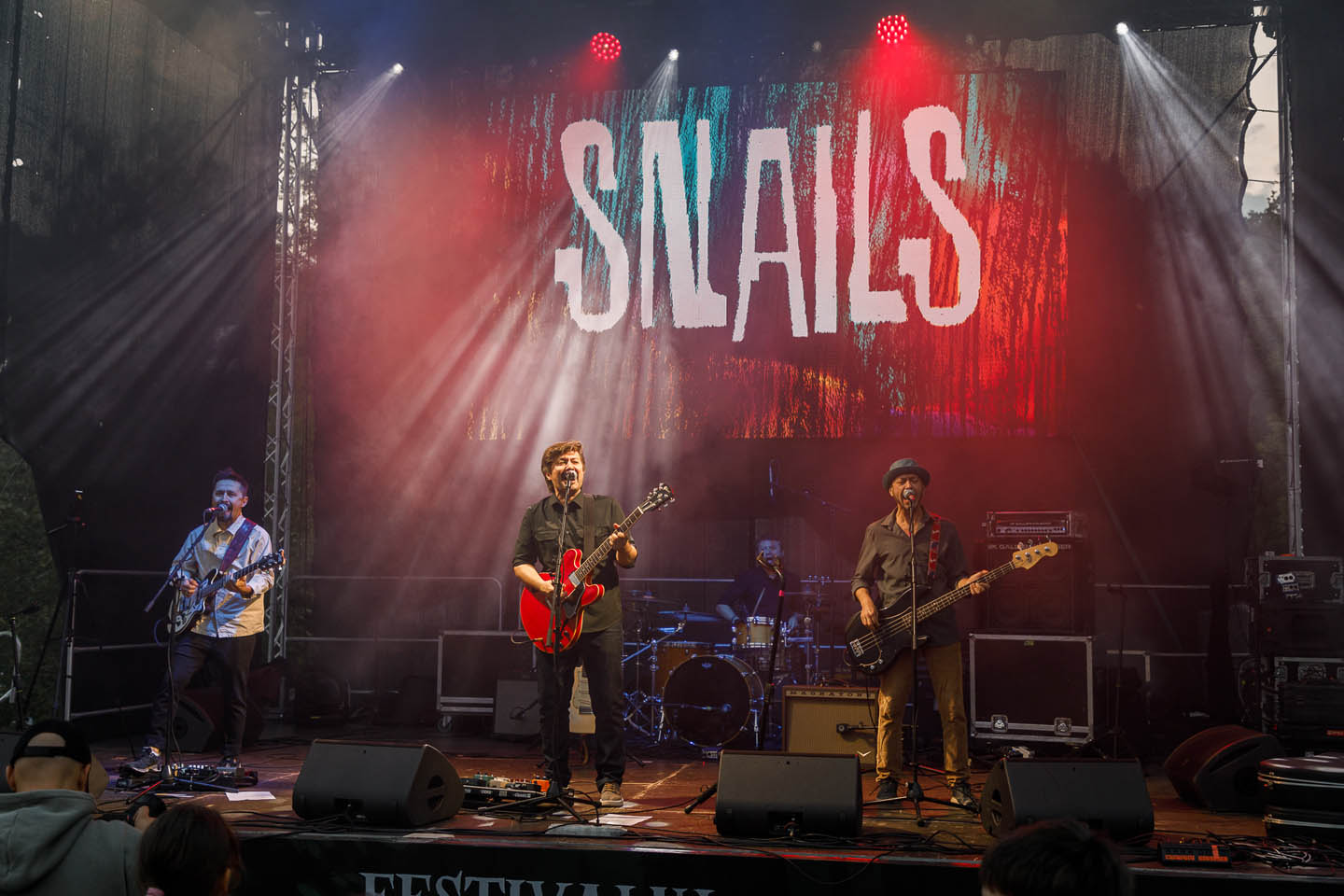 Музыкальная группа на сцене с гитарами и ударной установкой, над ними яркая вывеска с надписью SNAILS.