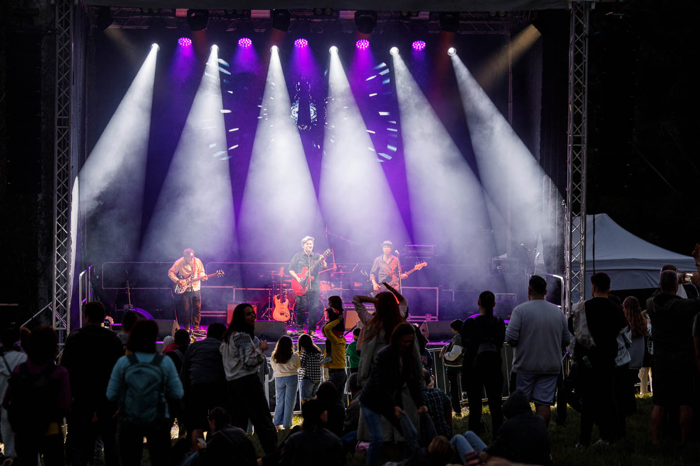 Музыкальная группа играет на сцене с яркими прожекторами, а перед сценой стоит и сидит аудитория.