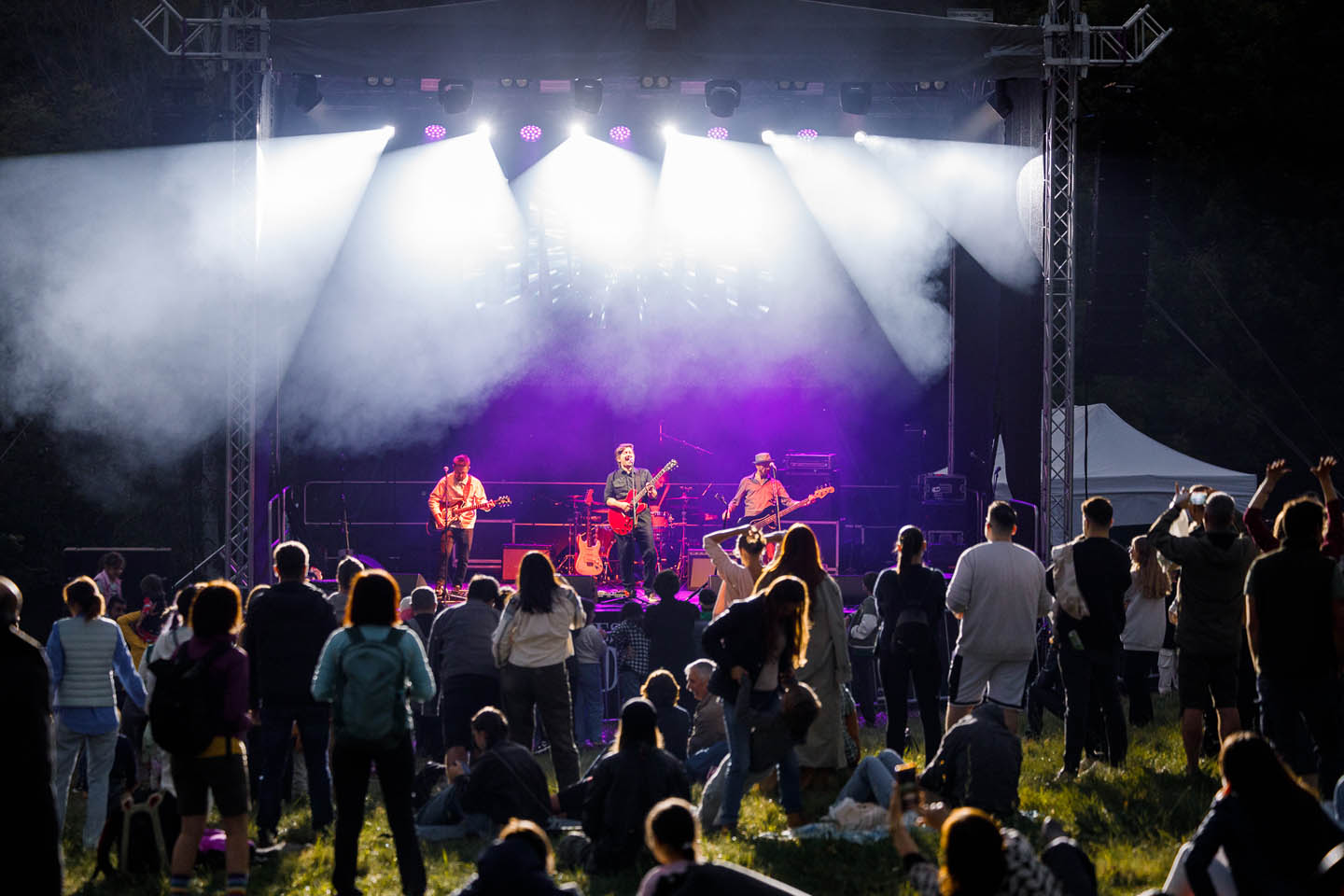 Концерт на открытом воздухе с группой из трех музыкантов на сцене и зрителями на траве, освещёнными белыми и фиолетовыми прожекторами.