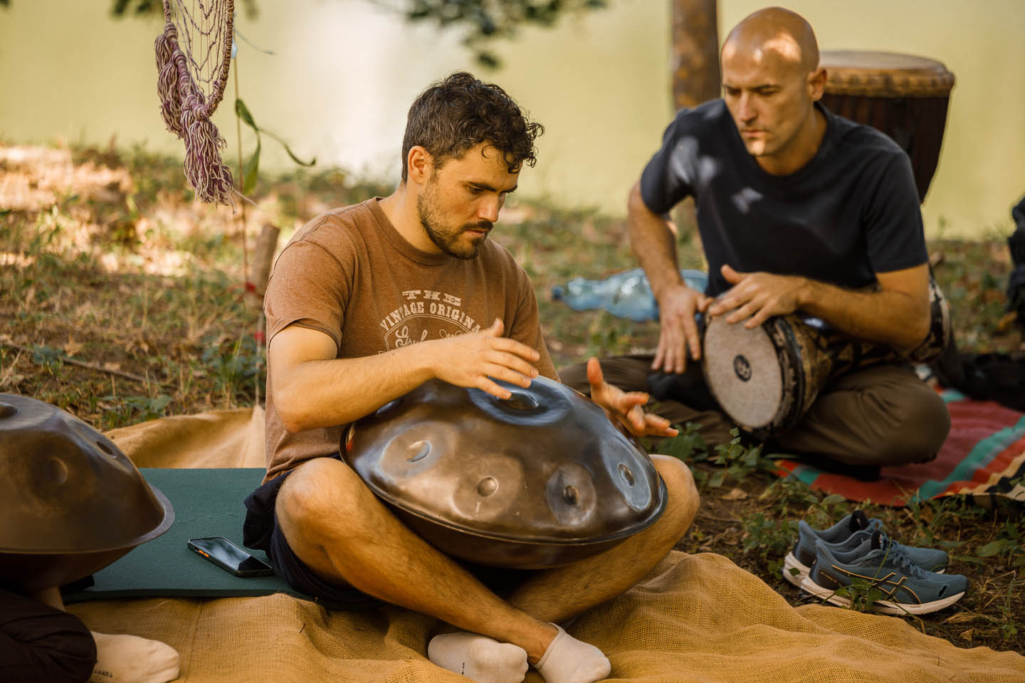 Два мужчины сидят на земле на пледах, играя на этнических перкуссионных инструментах на открытом воздухе.