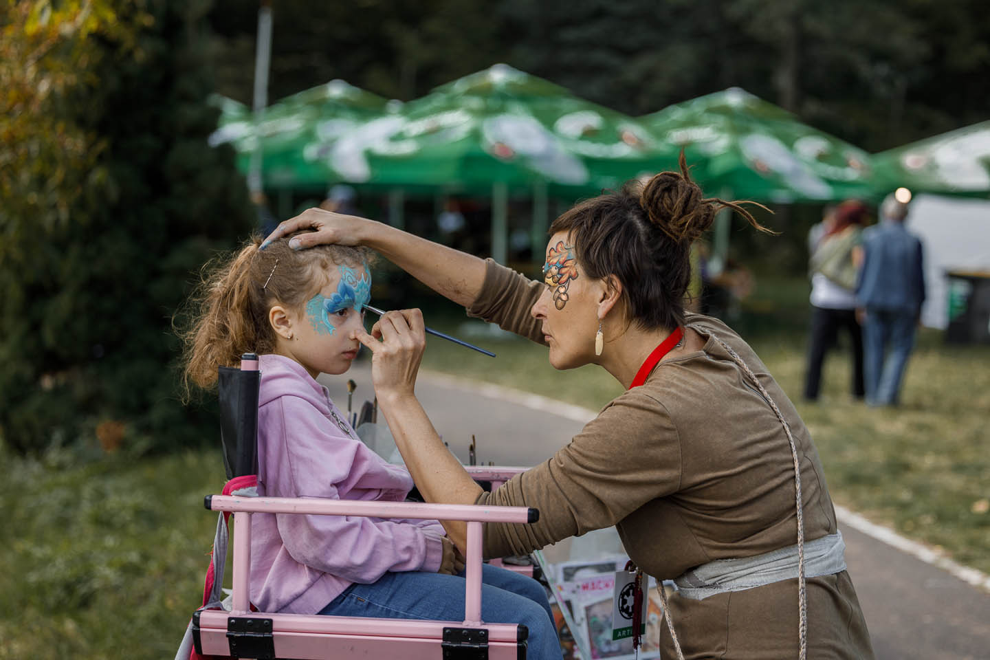 Женщина с раскрашенным лицом рисует узор на лице девочки в розовом свитере на улице.