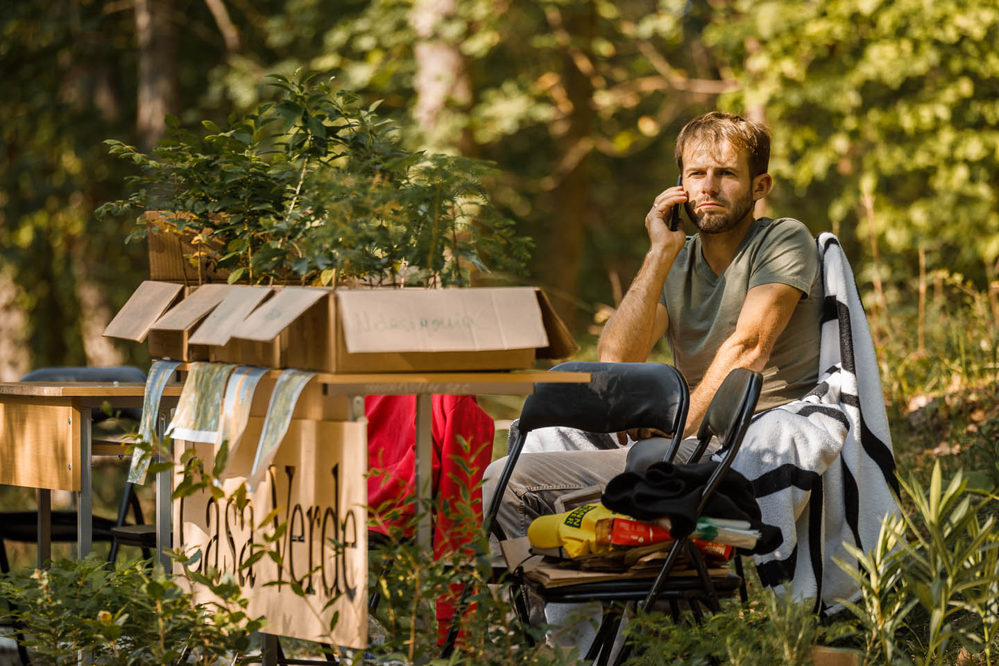 Мужчина разговаривает по мобильному телефону, сидя на стуле на улице рядом с коробками с саженцами растений.