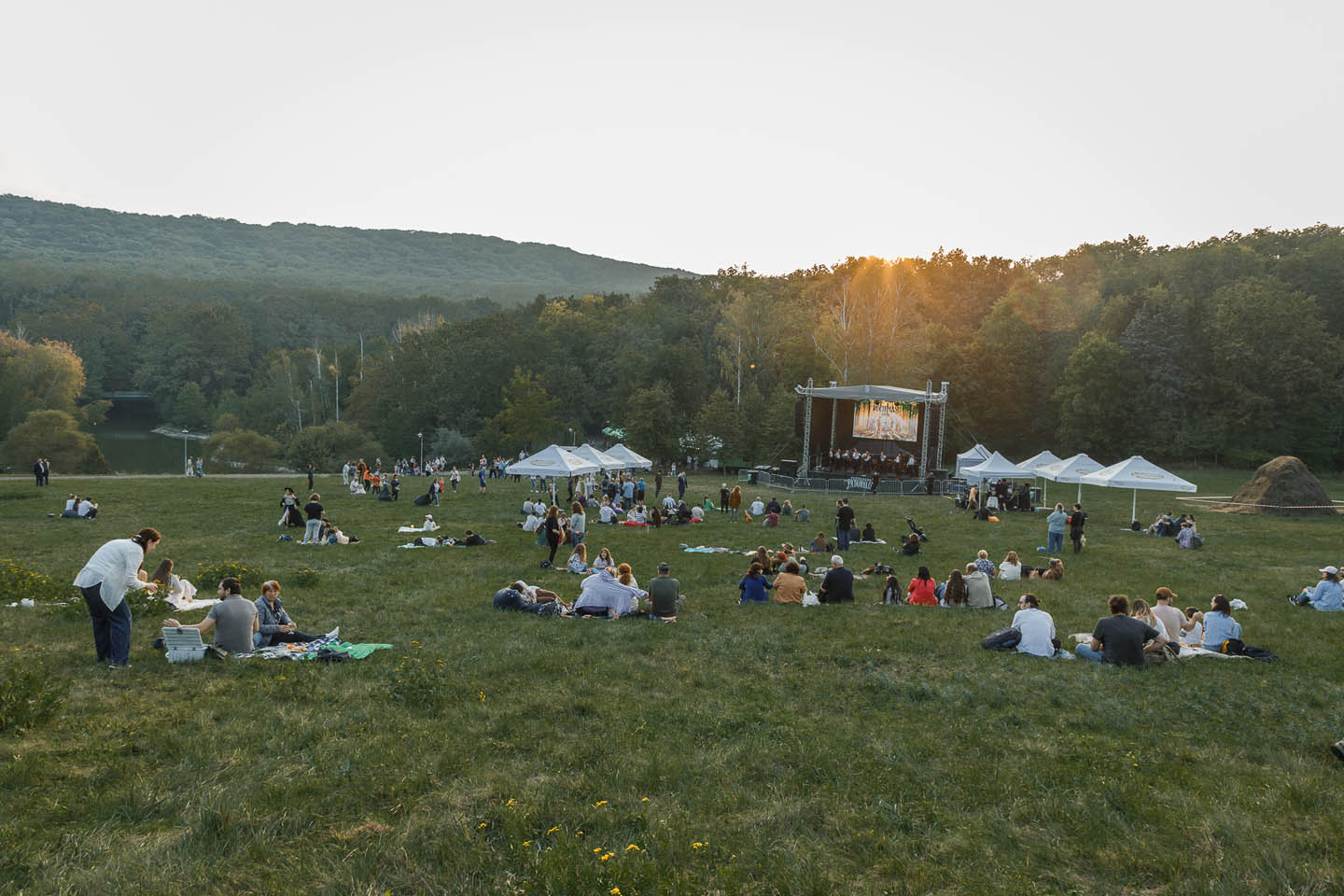 Люди на травянистом поле пикникуют и наслаждаются живым выступлением на сцене на фоне леса и холмов на закате.