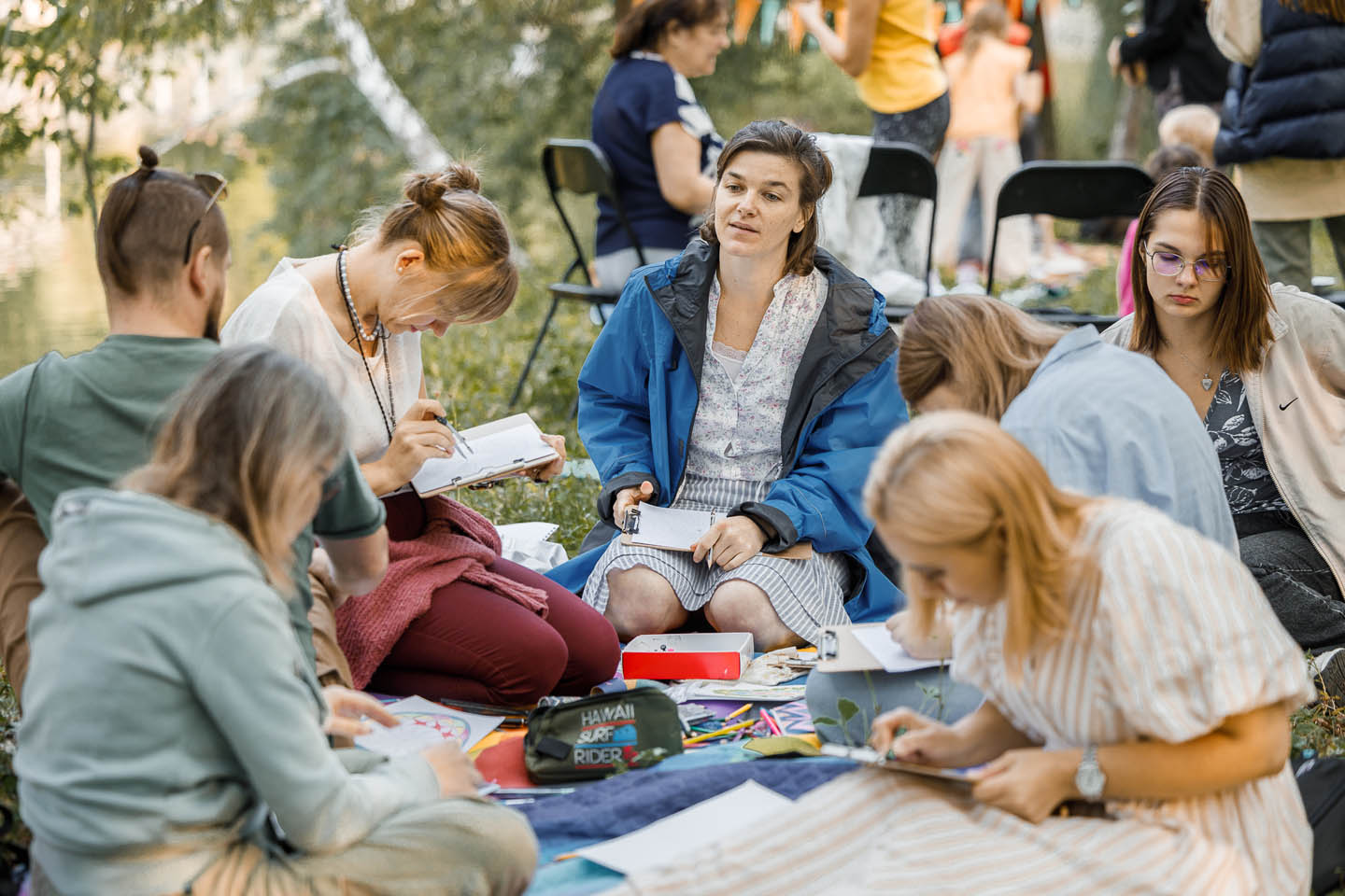 Группа людей занимается творчеством на открытом воздухе, рисуя и раскрашивая на листах бумаги в парке.