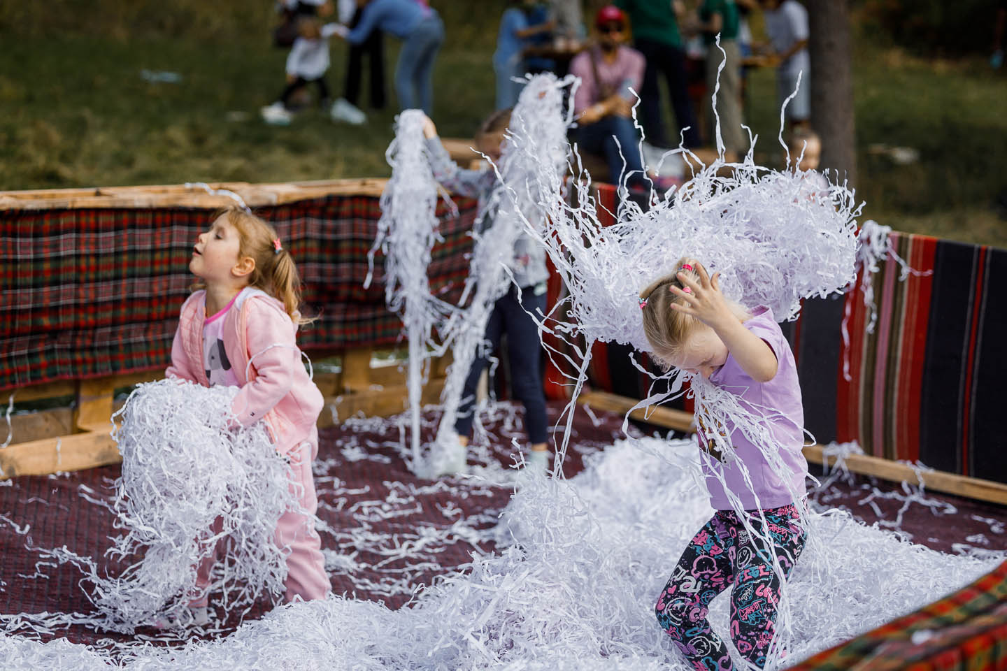 Дети играют с белыми бумажными нитями на открытом воздухе в огражденном пространстве с ковровым покрытием.