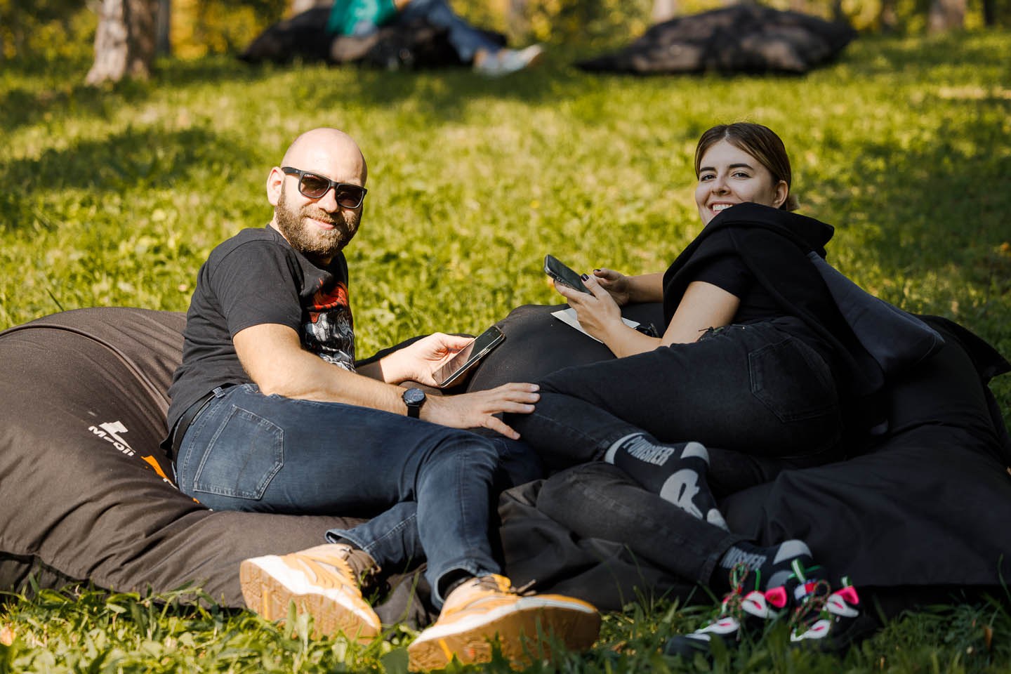 Мужчина в солнцезащитных очках и женщина улыбаются, лежа на черных надувных креслах на траве в солнечный день.