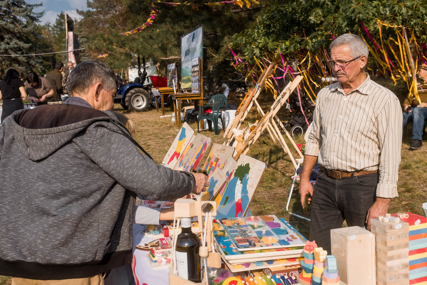 Мужчина покупает образовательные деревянные игрушки с картами, а продавец стоит рядом на уличной ярмарке с разноцветными лентами вокруг.