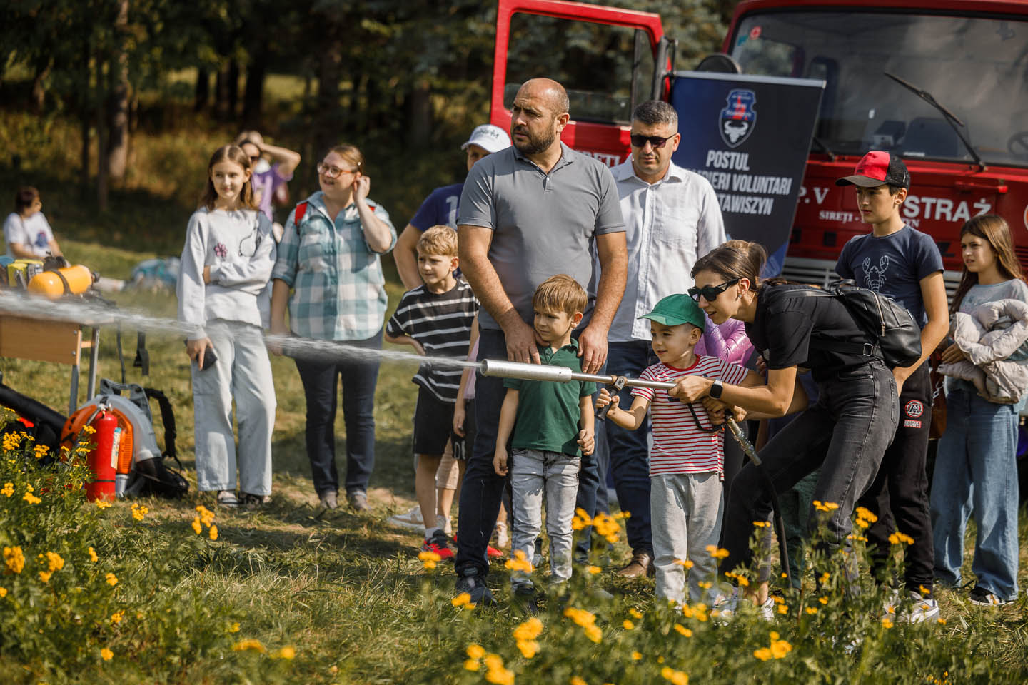Женщина в очках помогает мальчику направить пожарный шланг, из которого бьет струя воды, на фоне группы детей и взрослых у красной пожарной машины.