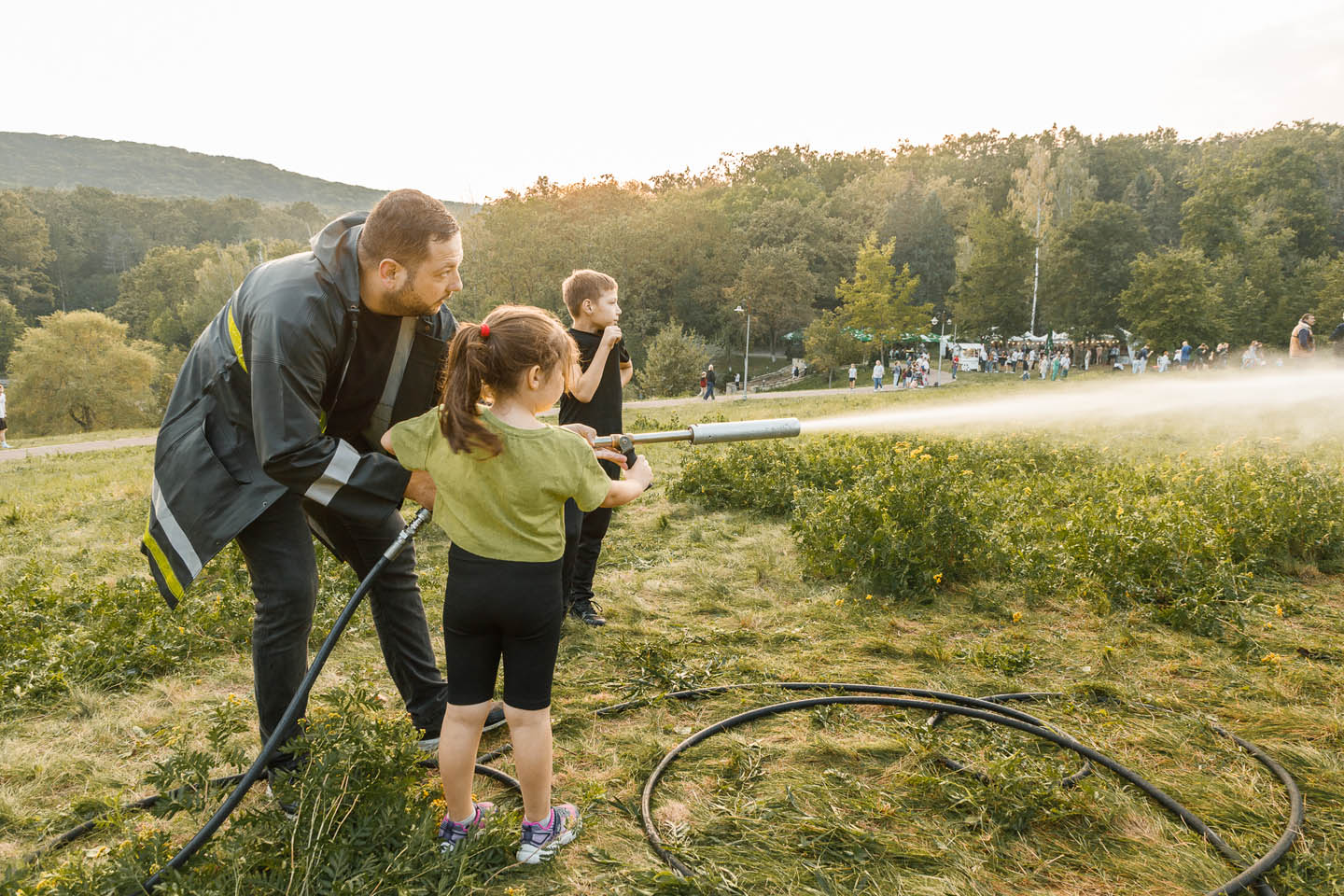 Пожарный обучает девочку использовать водяной шланг в парке, на фоне мальчик и группа людей среди деревьев.