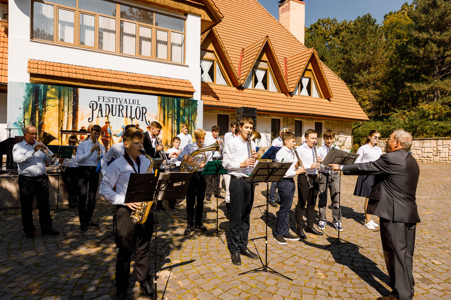 Музыкальная группа молодых людей играет на духовых инструментах под руководством дирижера на открытом воздухе возле дома с оранжевой крышей.