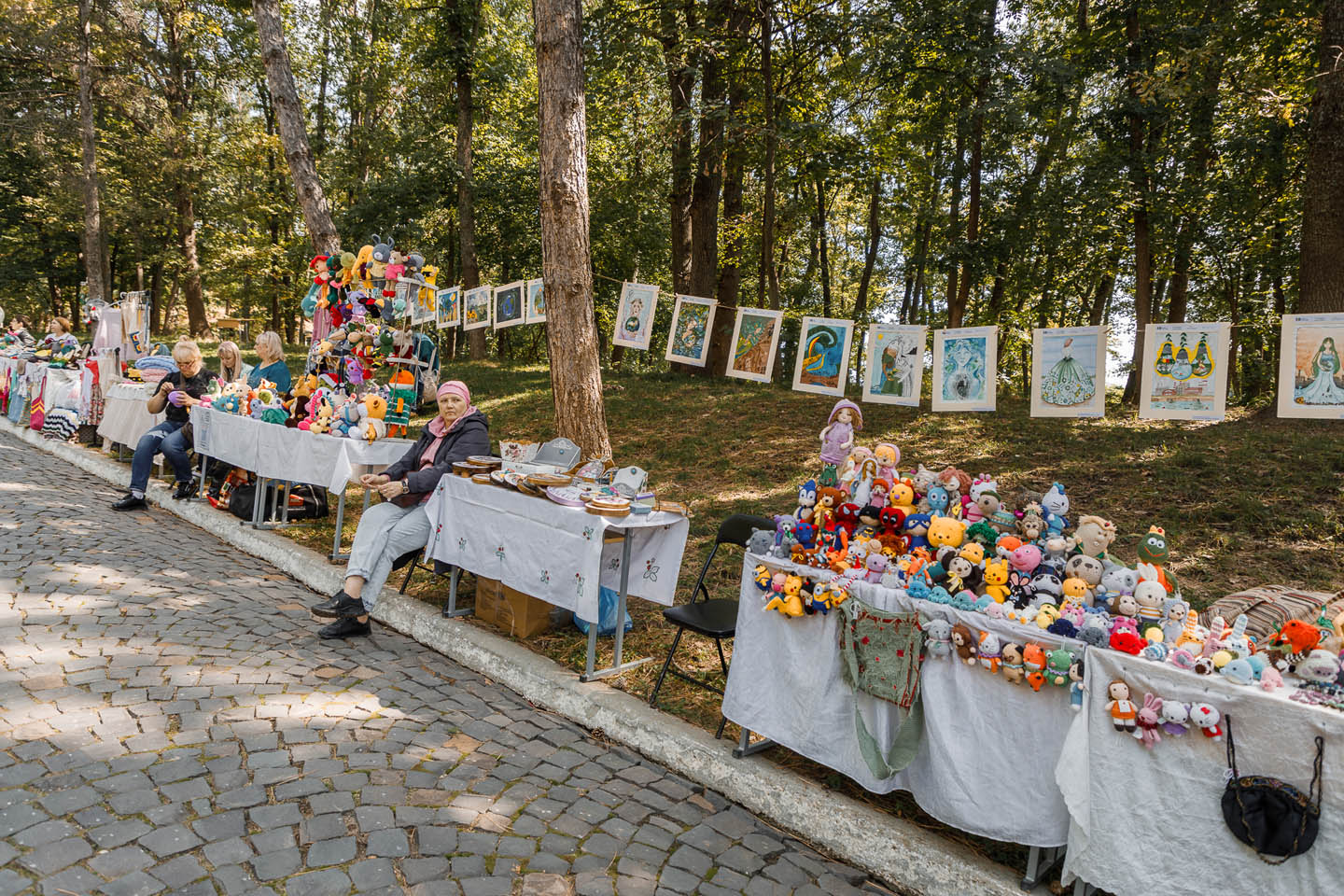 Женщины за столами с разнообразными вязанными игрушками и картинами, развешанными на деревьях в парке.