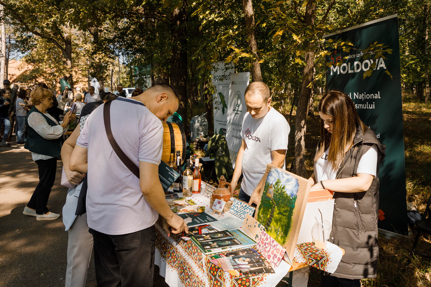 Люди рассматривают товары на столе с традиционной скатертью на открытом воздухе в парке, рядом с баннерами с надписью Молдова.