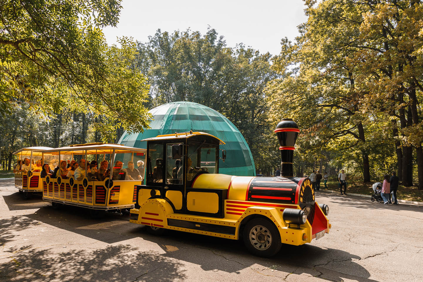 Ярко-желтый мини-паровой туристический поезд с пассажирами на фоне зеленого купола и деревьев в парке.