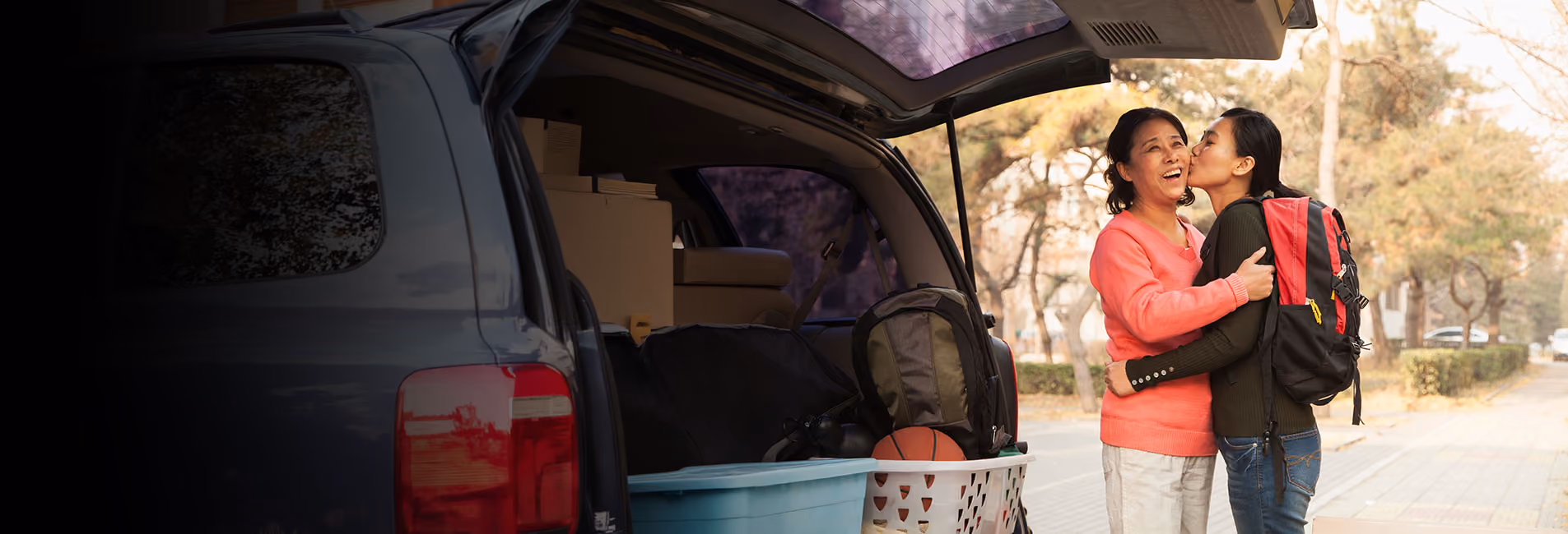 Young woman hugs mom while standing by car loaded up for move to college.