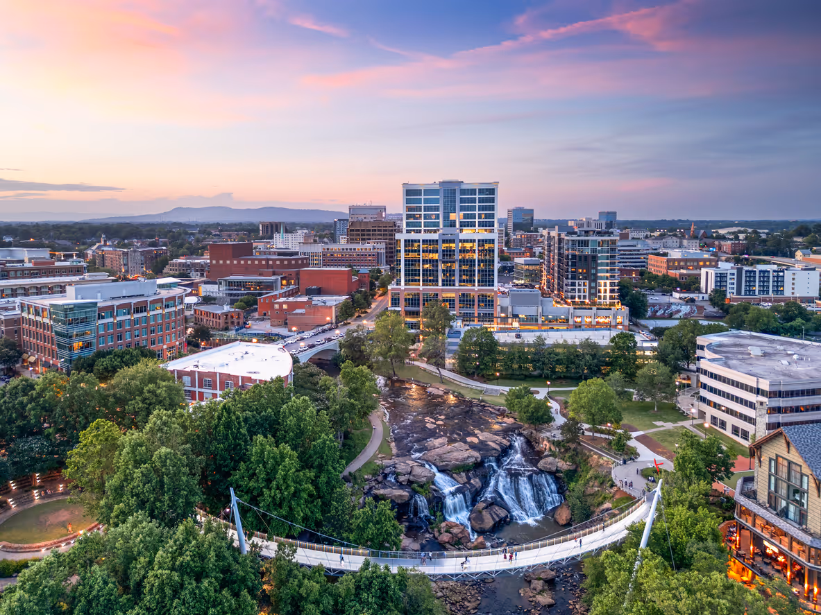 Cityscape at sunset showing a river with waterfalls, a pedestrian suspension bridge, downtown buildings, and trees.