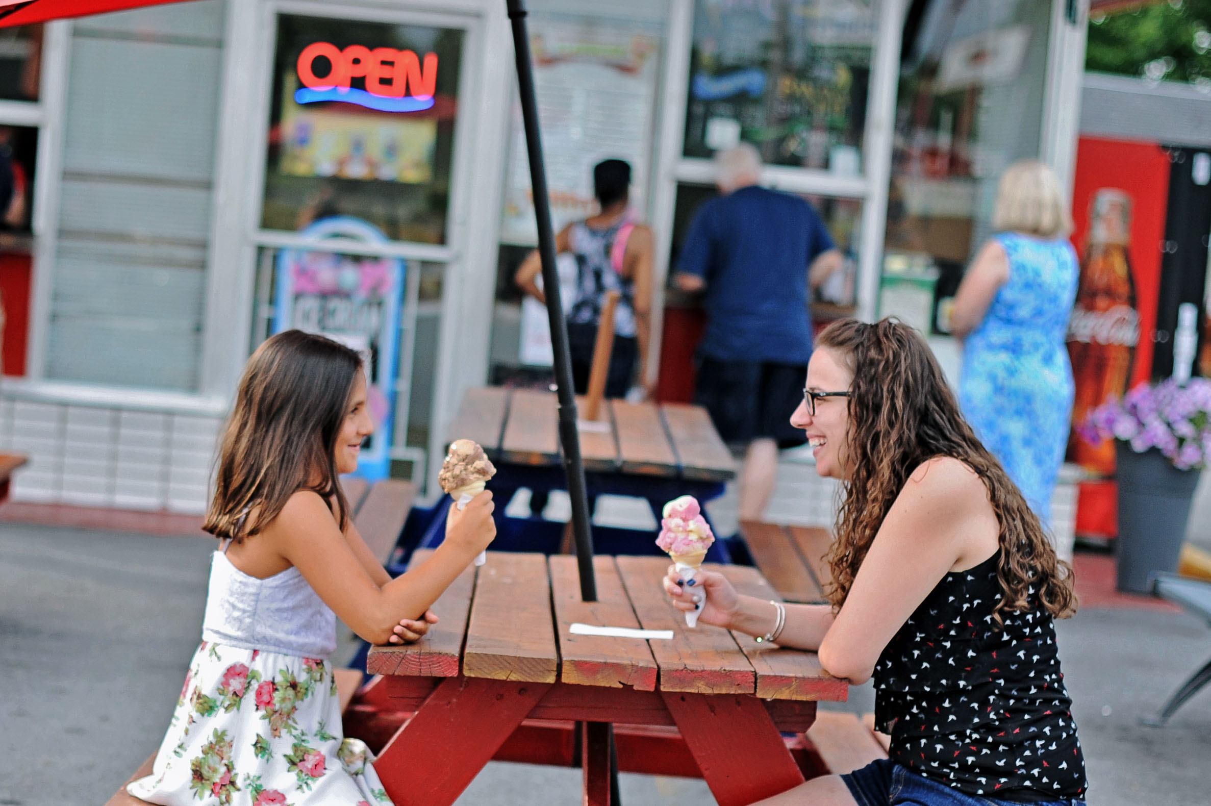Two girls sitting at a wooden picnic table, smiling and holding ice cream cones outside an ice cream shop with an open sign.