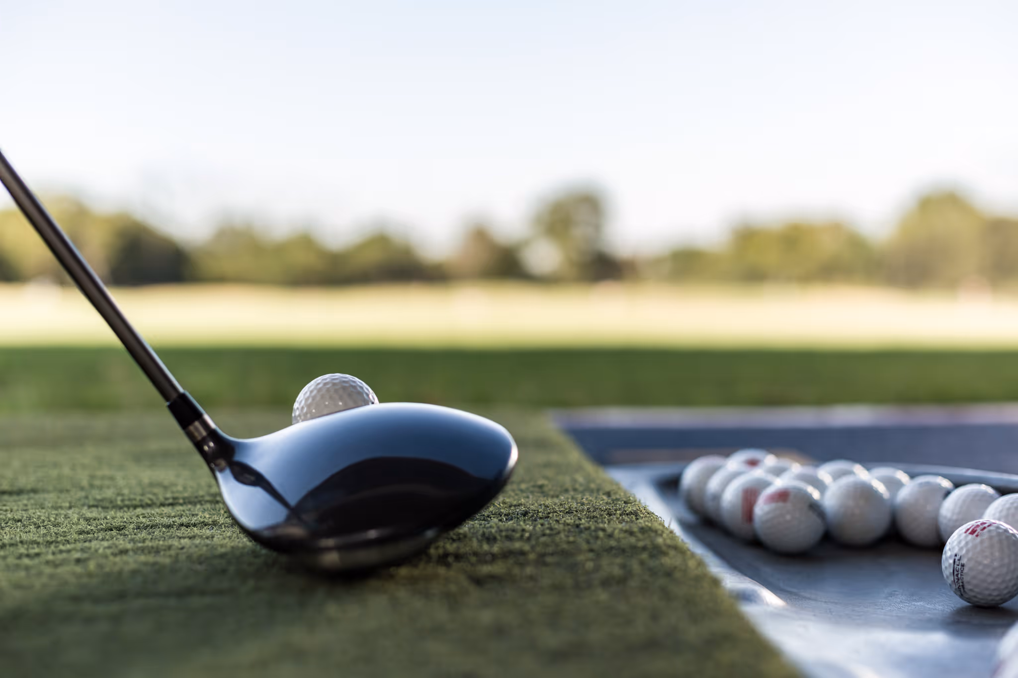 Close-up of a golf driver poised behind a golf ball on artificial turf, with a tray of golf balls in the background.