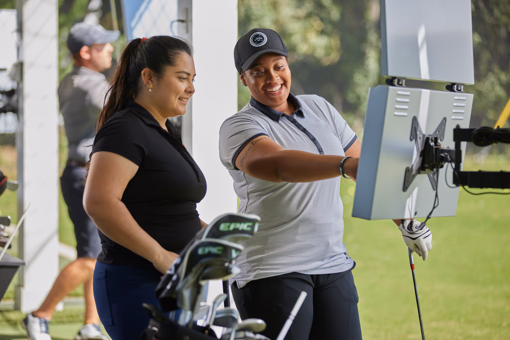 Two women smiling and looking at a screen at a golf driving range, with a man practicing golf swing in the background.