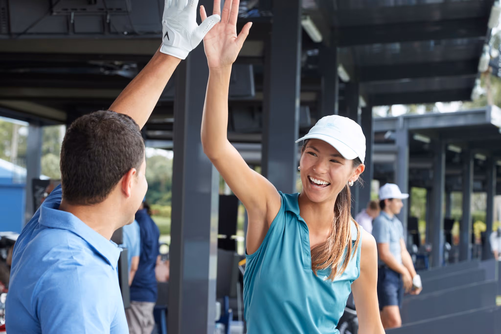 Smiling young woman in a teal sleeveless golf shirt and white cap giving a high five to a man in a blue shirt at a golf driving range.