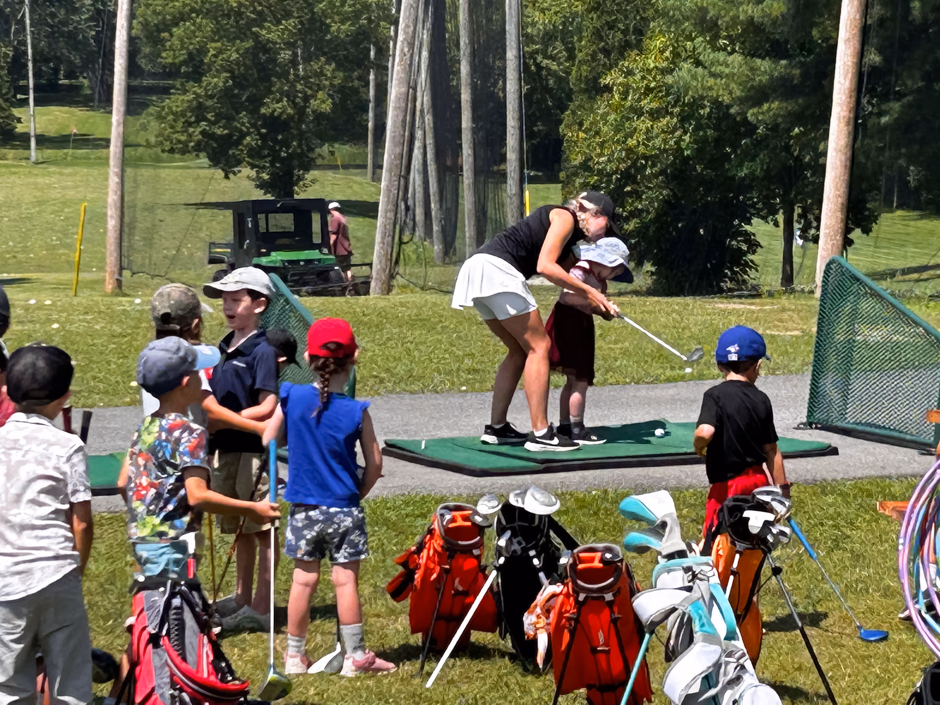 Golf instructor teaching a young child to swing a golf club at Archie's driving range with other children and golf bags nearby.