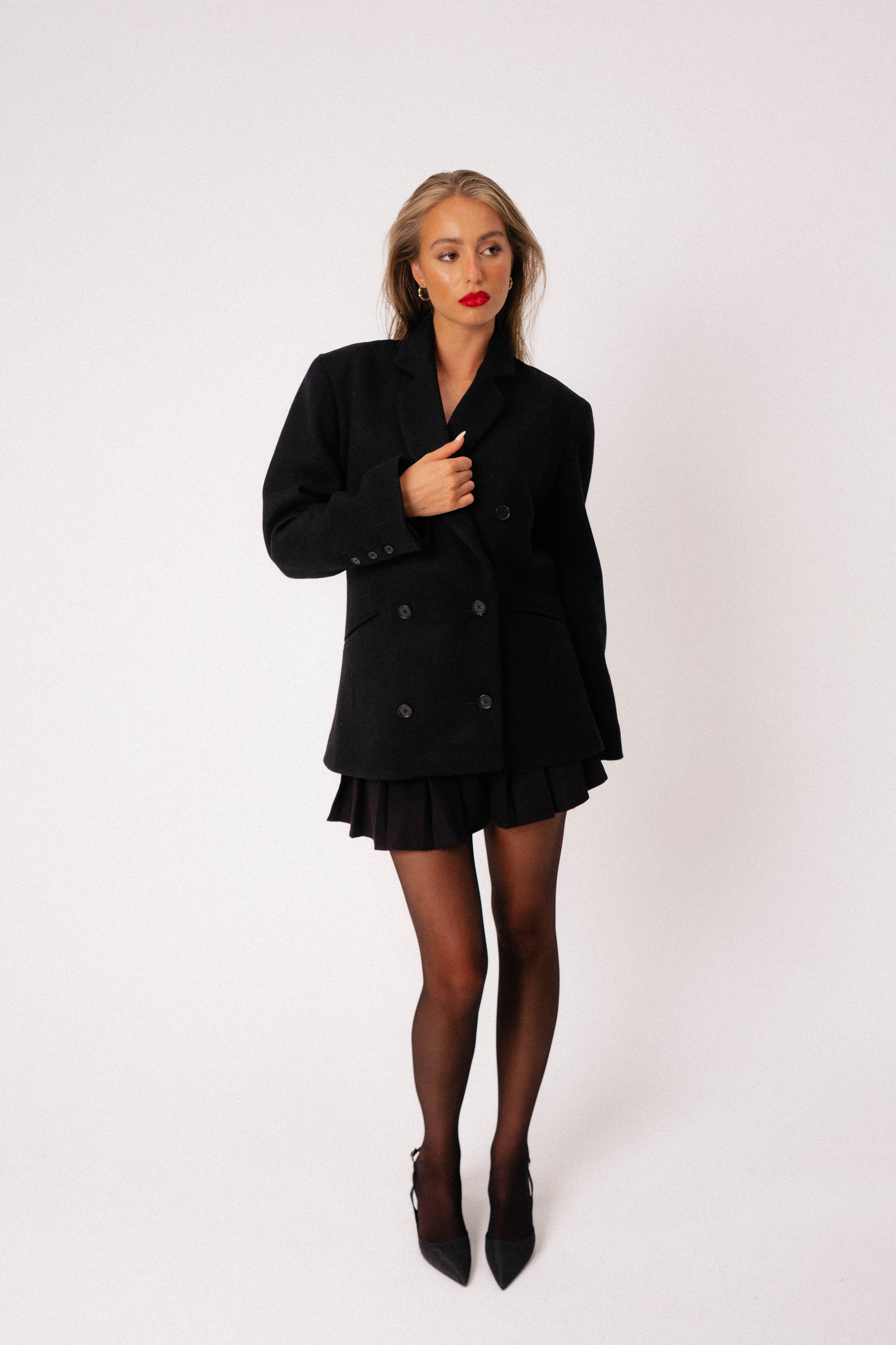 A model in a studio wearing a black coat by Lutea Studios dressed in a skirt.