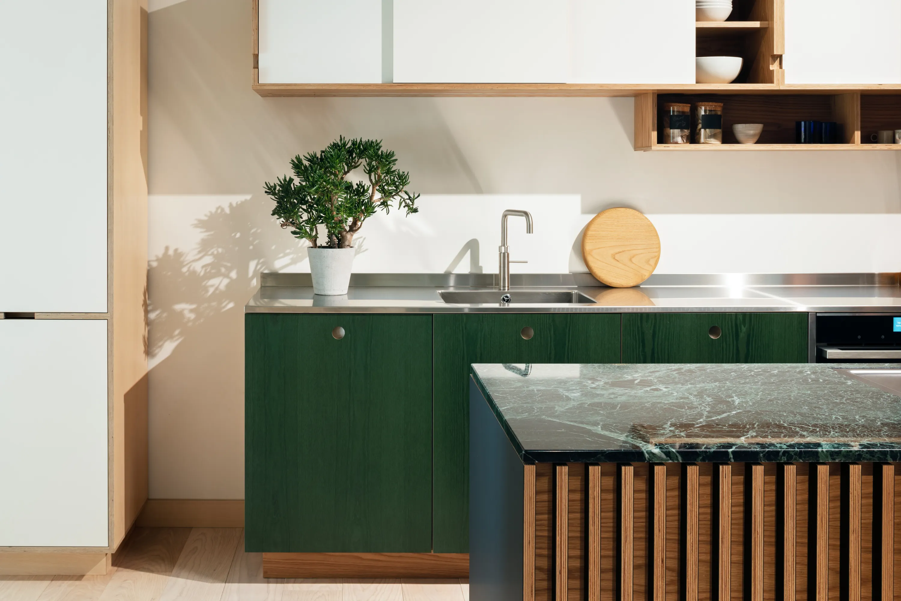 Modern kitchen with green lower cabinets, stainless steel countertop and sink, potted plant, wooden cutting board, and marble kitchen island.
