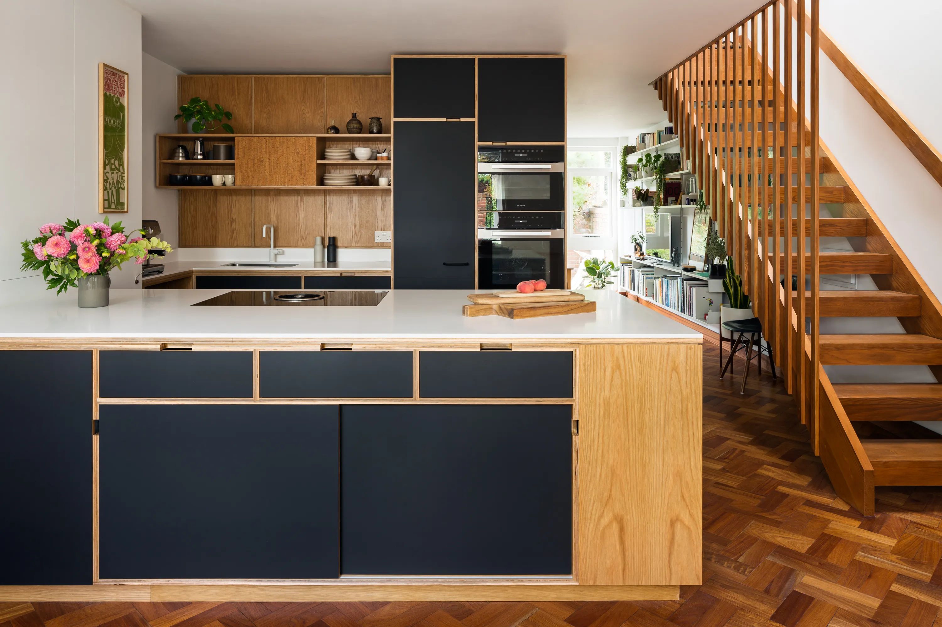 Contemporary oak-veneered plywood kitchen with navy cabinetry, white island and timber staircase by Uncommon Projects