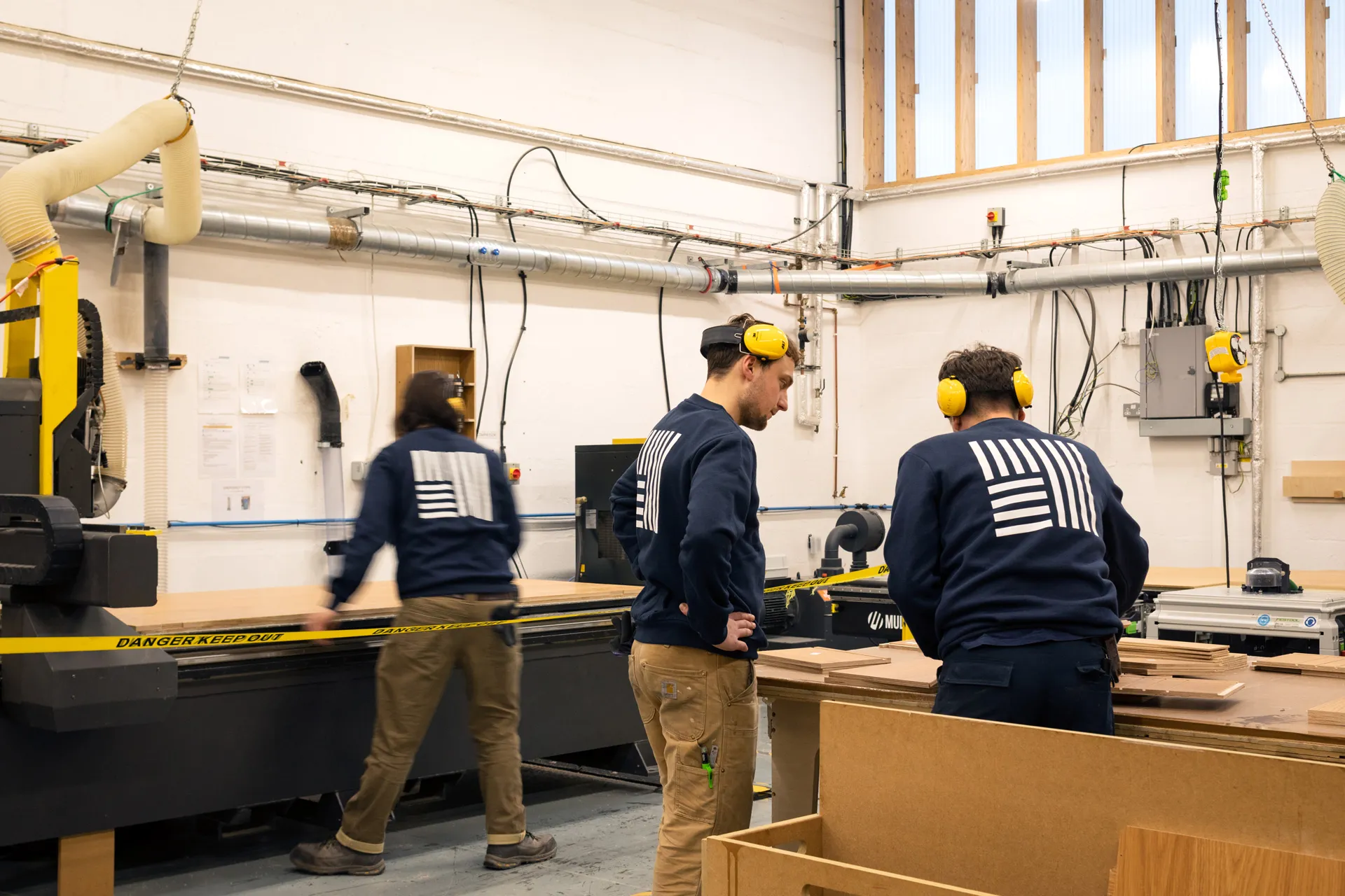 Three people wearing matching navy sweatshirts and yellow ear protection working in a woodworking workshop with large machines and wood panels.