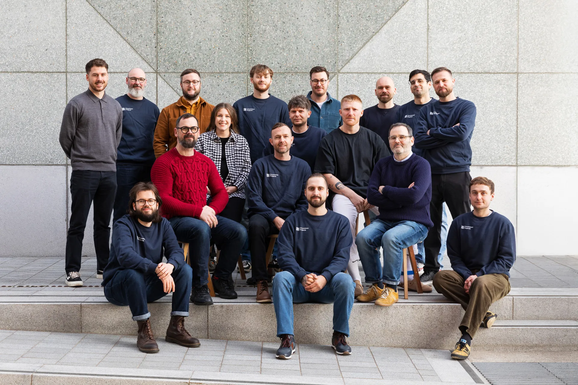 Group portrait of seventeen casually dressed people sitting and standing in front of a tiled wall outdoors.