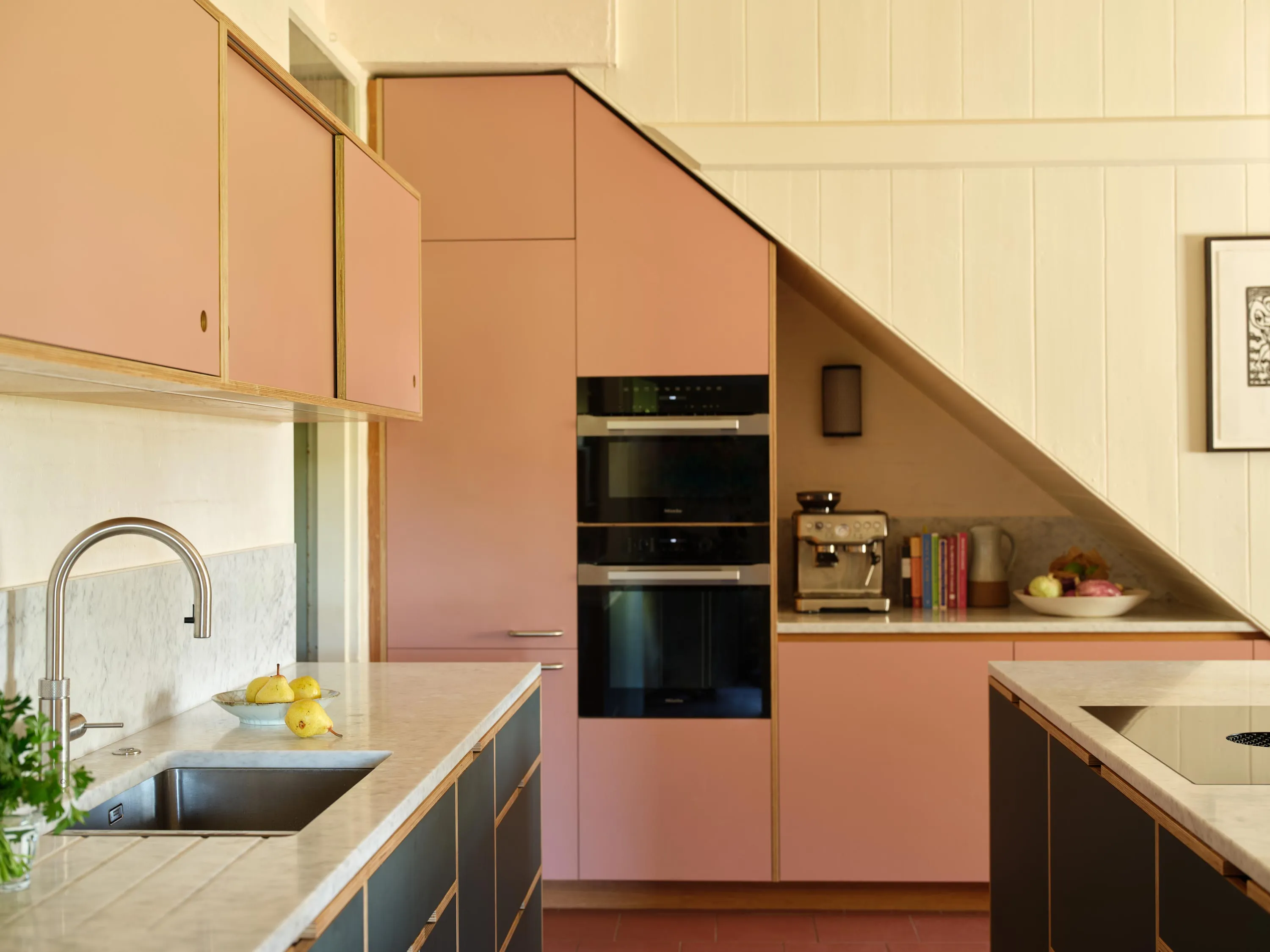 Modern kitchen with pink cabinets and black lower drawers, a marble countertop, built-in ovens, a coffee machine, cooktop, and a stainless steel sink with a curved faucet.