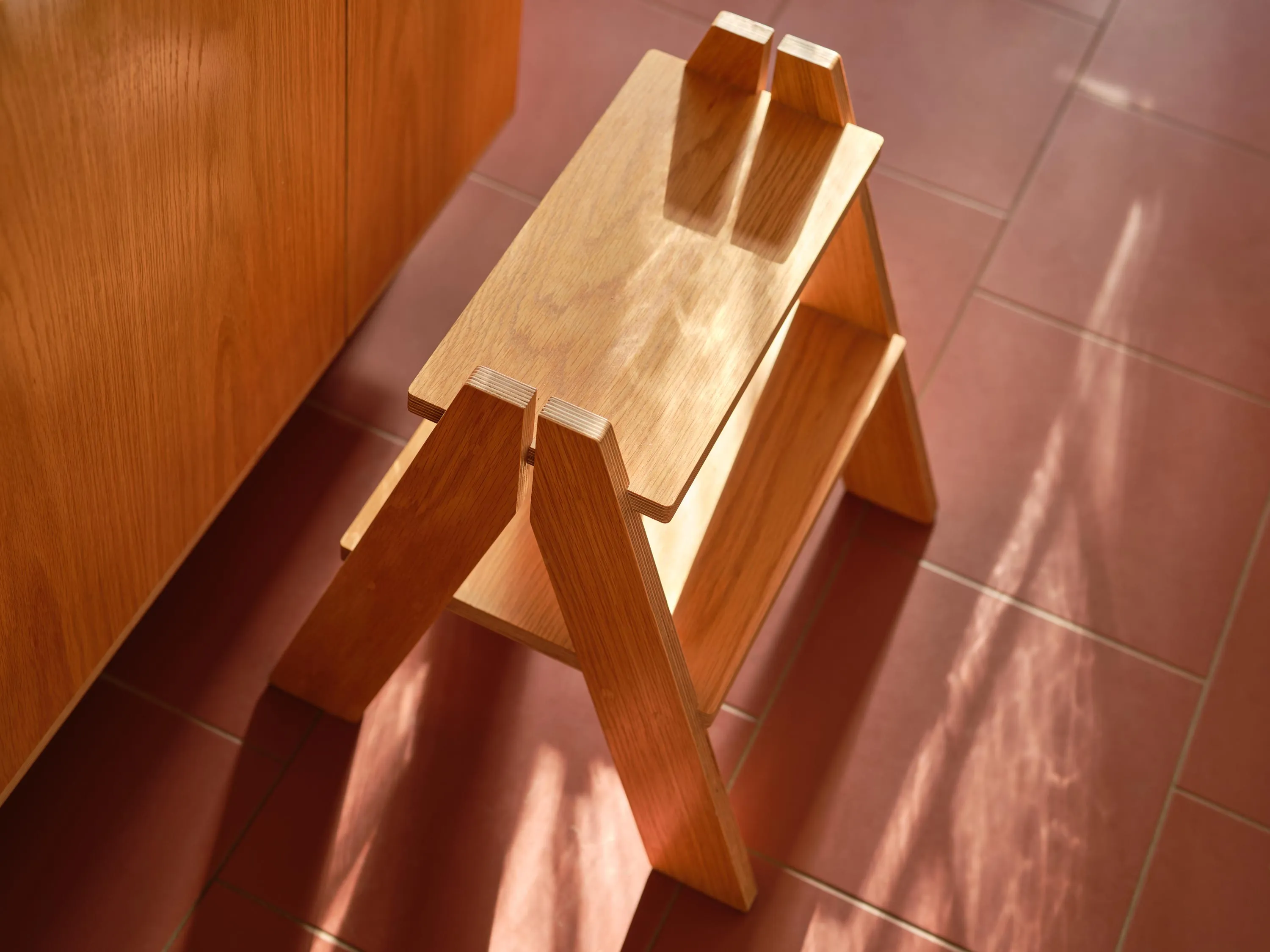 Small wooden step stool with two platforms on a red tiled floor next to wooden cabinetry.
